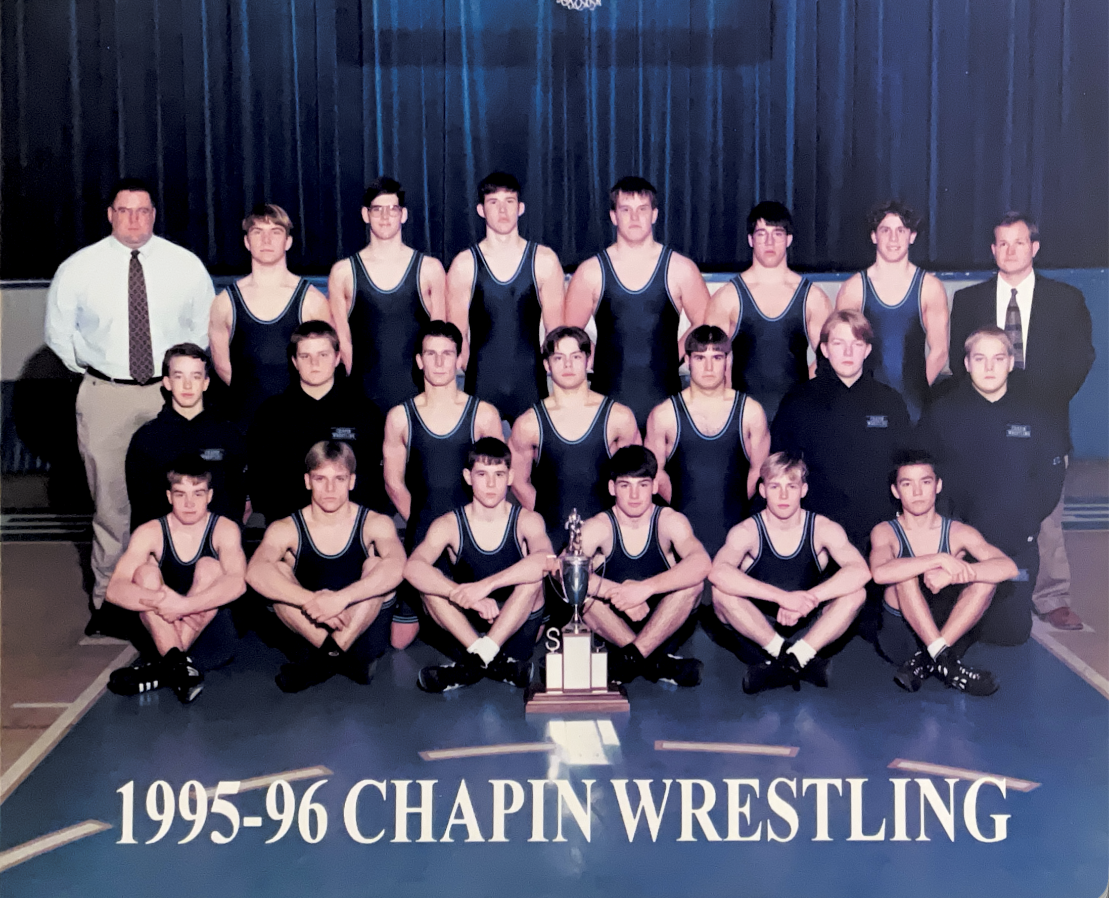 A wrestling team from 1995-96, posing with a trophy in front of a black curtain. The team members are in navy blue singlets, with two coaches and three staff members standing behind. The banner at the bottom reads "1995-96 Chapin Wrestling."