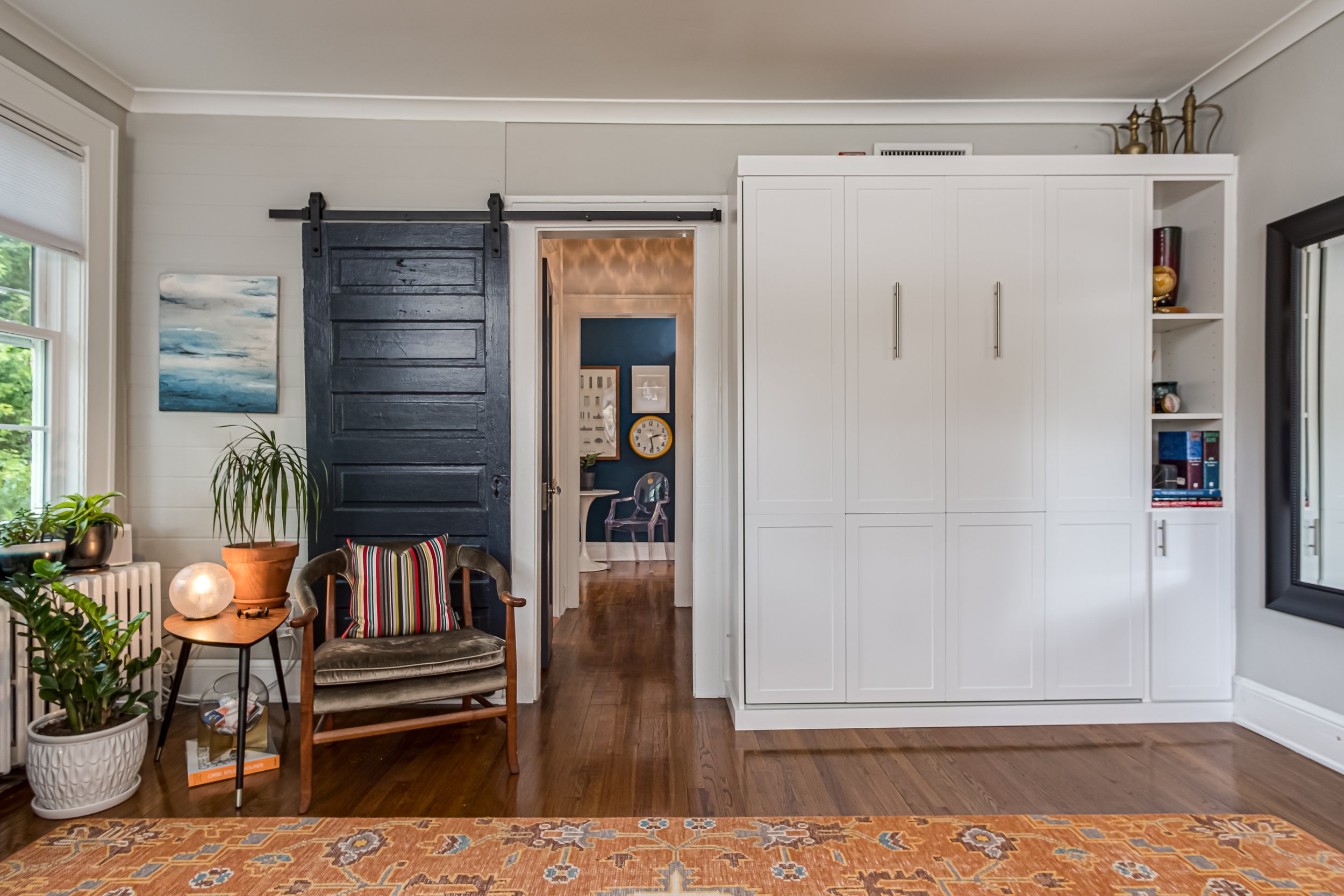 Interior room with a dark sliding barn door, white murphy bed with storage cabinetry, a wooden chair with a striped pillow, a side table with a lamp and plant, and visible hallway.