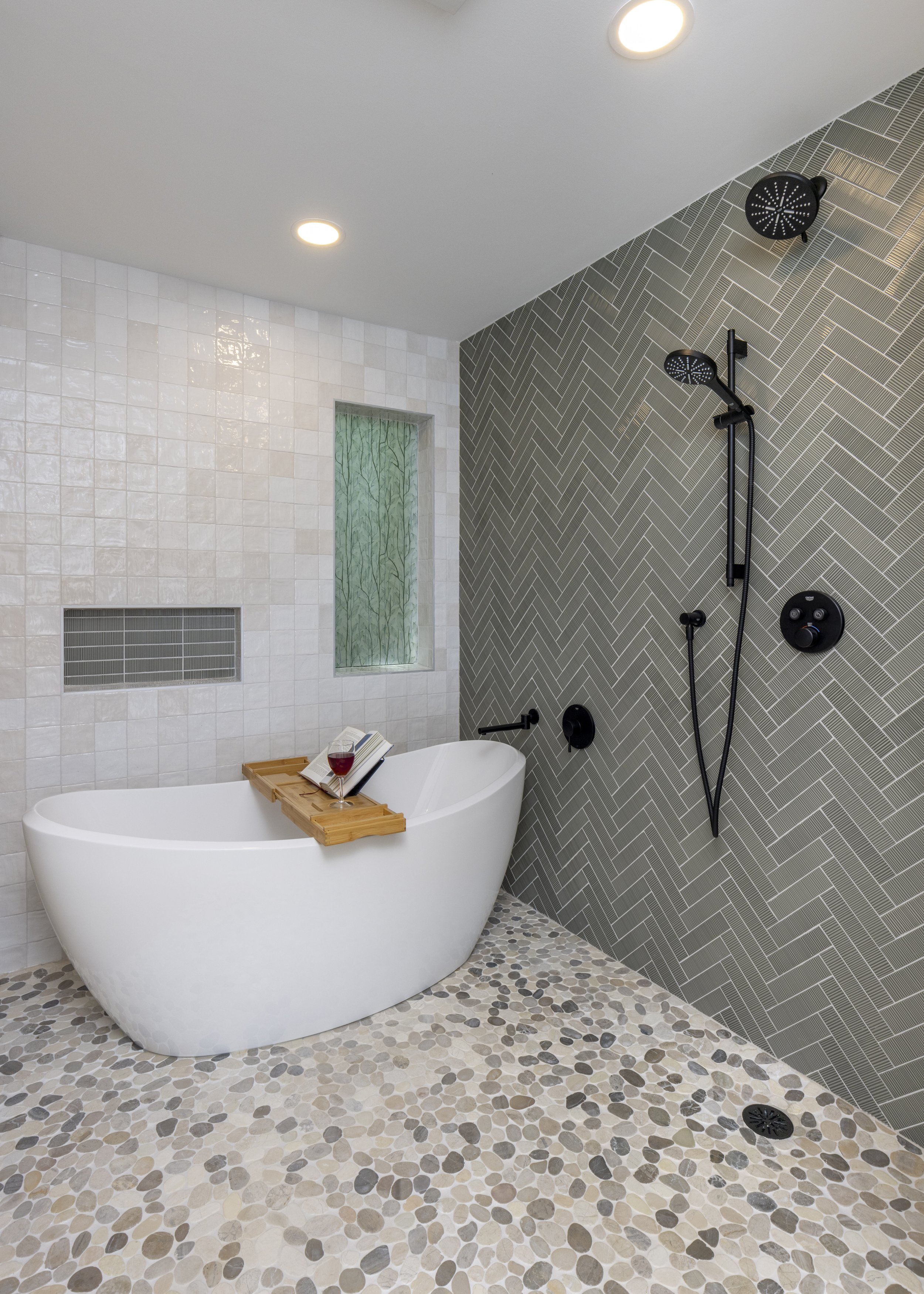 Modern bathroom with freestanding white bathtub, wooden bath tray holding a book and glass of red wine, herringbone-pattern tiled wall, black shower fixtures, and pebble stone flooring.