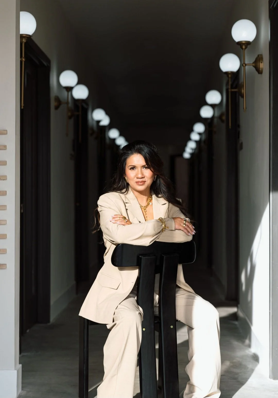 A woman sitting on a black chair in a hallway with modern wall sconces, wearing a beige suit and gold jewelry, with crossed arms and a confident expression.