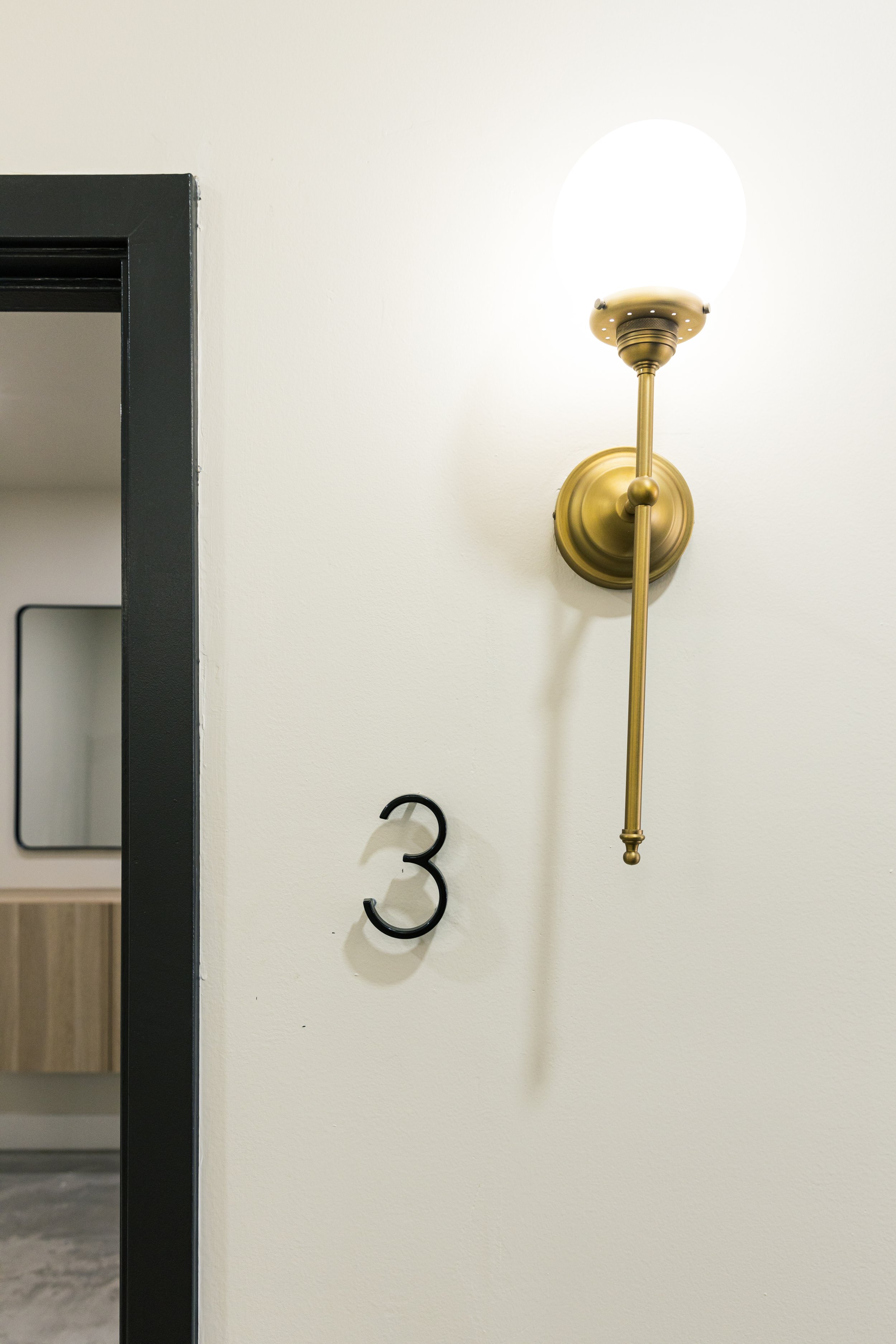 Wall with a black number 3 and a brass wall-mounted light fixture turned on, casting a shadow.