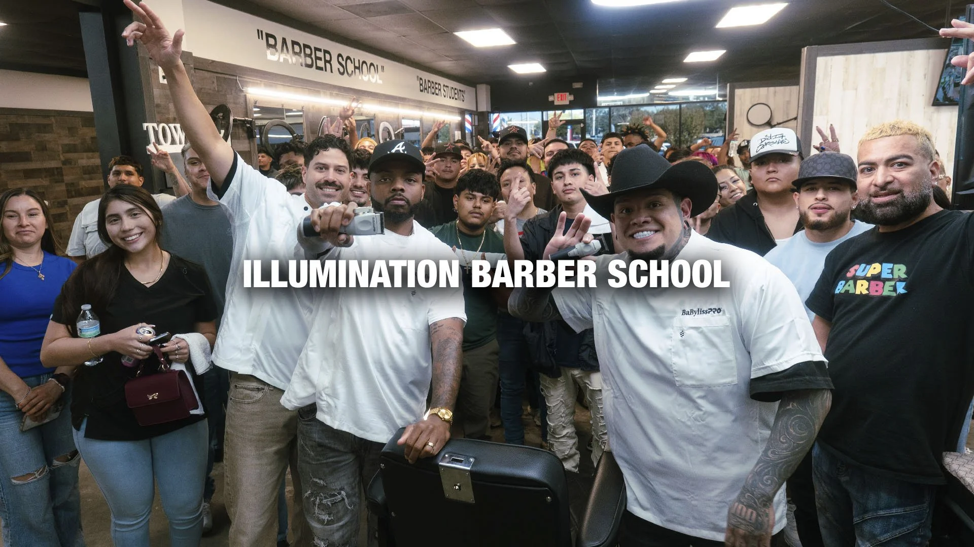 Group of people inside a barber school, with two men in the foreground wearing barber uniforms, one holding a camera. The background features a sign reading 'ILLUMINATION BARBER SCHOOL' and a crowd of students and staff.