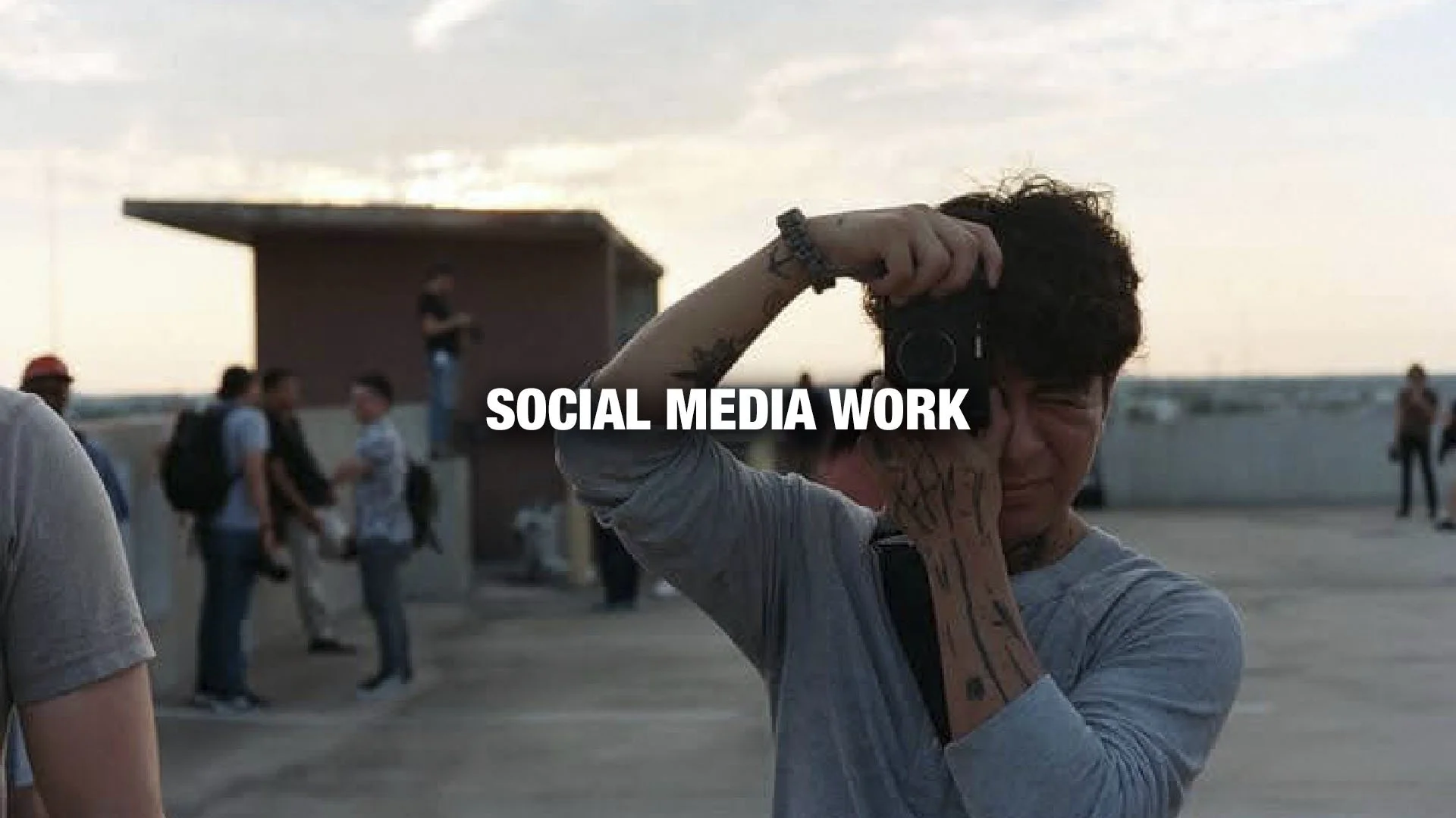 A person with curly hair taking a photograph on a rooftop at sunset, with a group of people in the background and the text 'Social Media Work' overlayed.