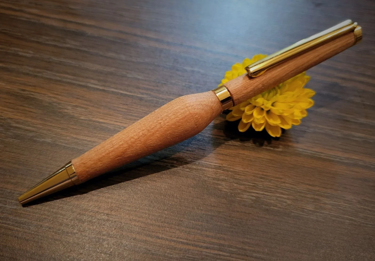 A pen with a wooden body and gold accents resting on a yellow flower on a wooden table.