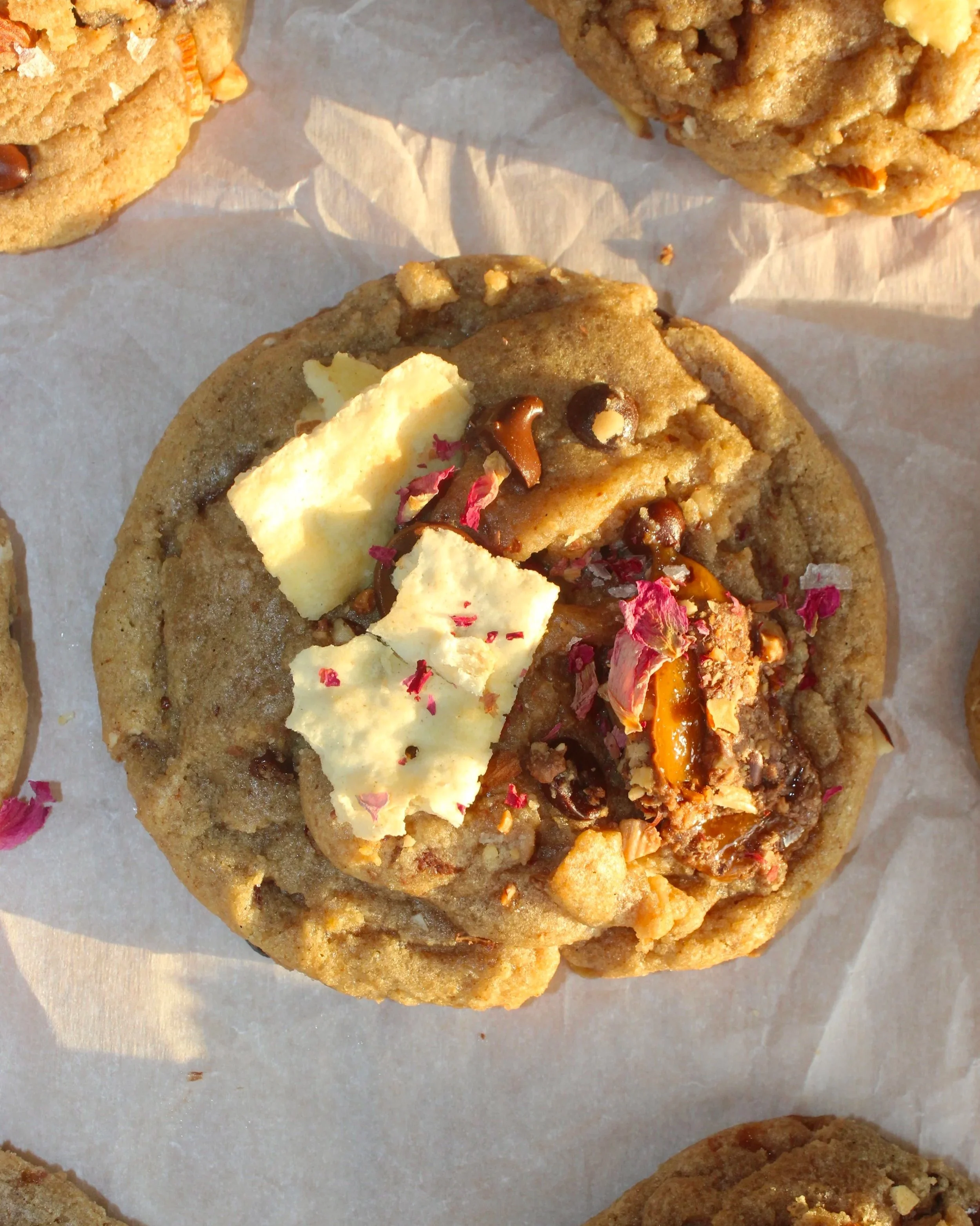Close-up of a freshly baked chocolate chip cookie topped with white chocolate chunks, fuchsia flower petals, and caramel crumbs on a parchment paper