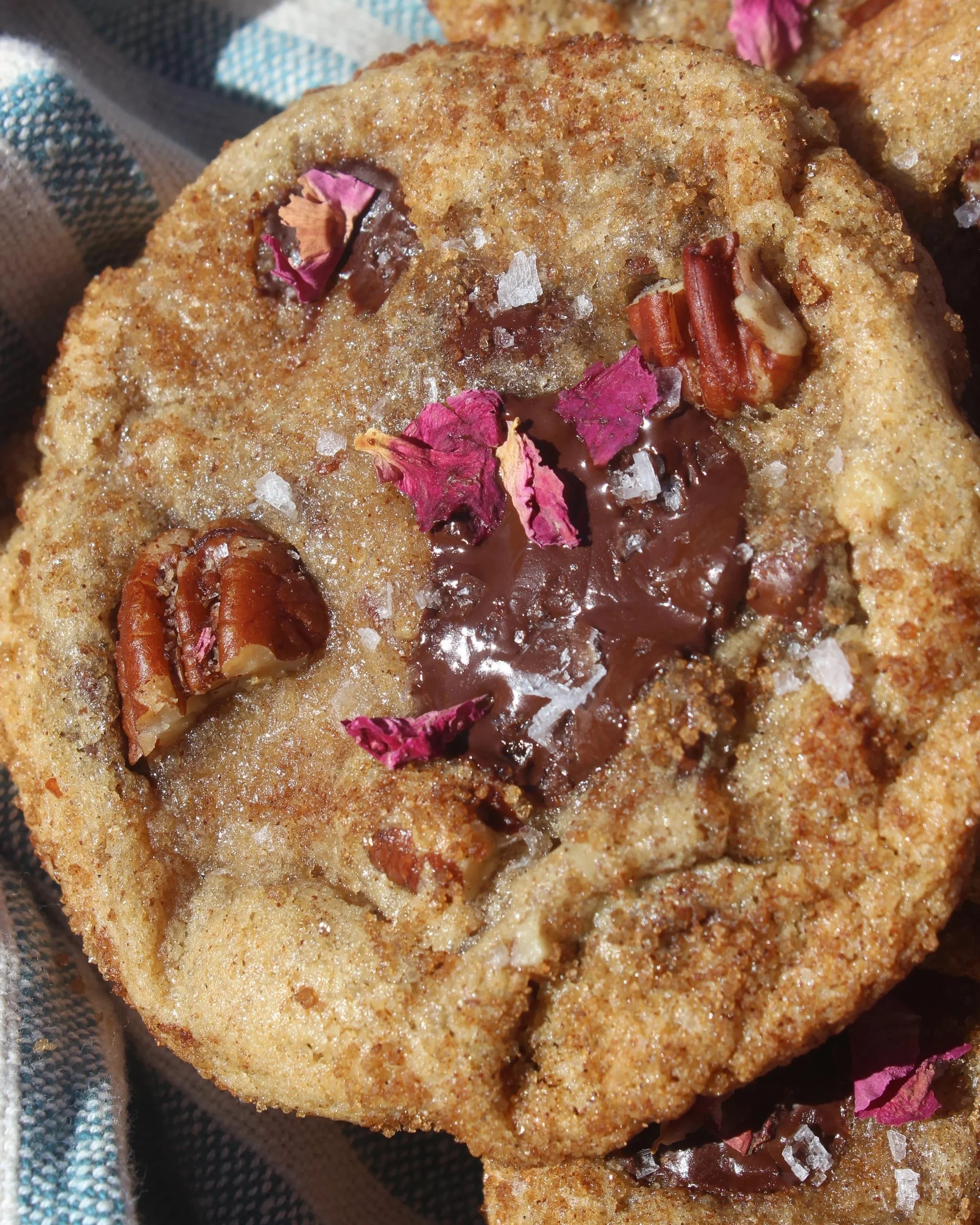 A close-up of a homemade cookie topped with melted chocolate, pink rose petals, chopped pecans, and sea salt flakes.
