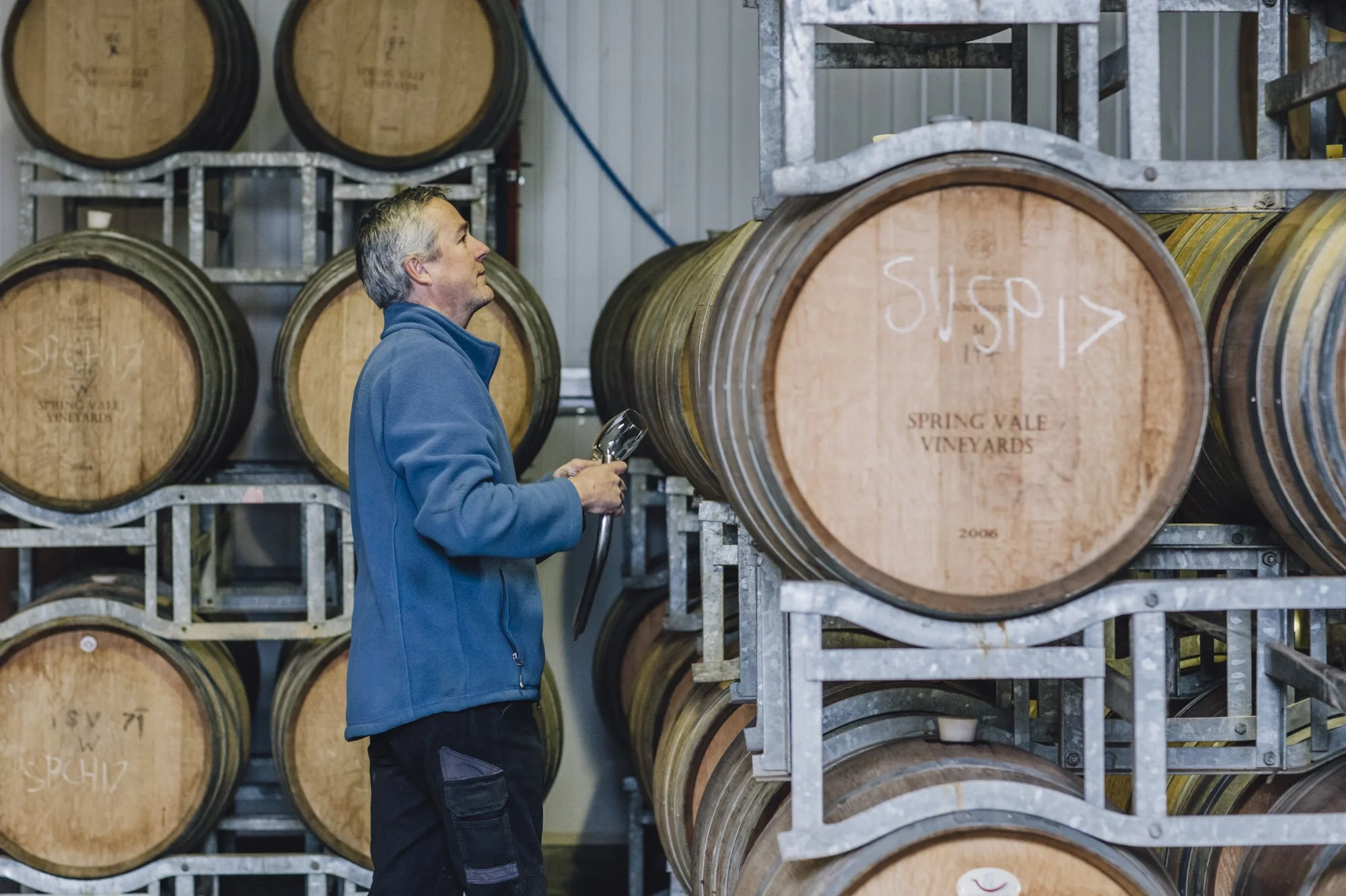 Man inspecting wine barrels in a winery, wearing a blue jacket.