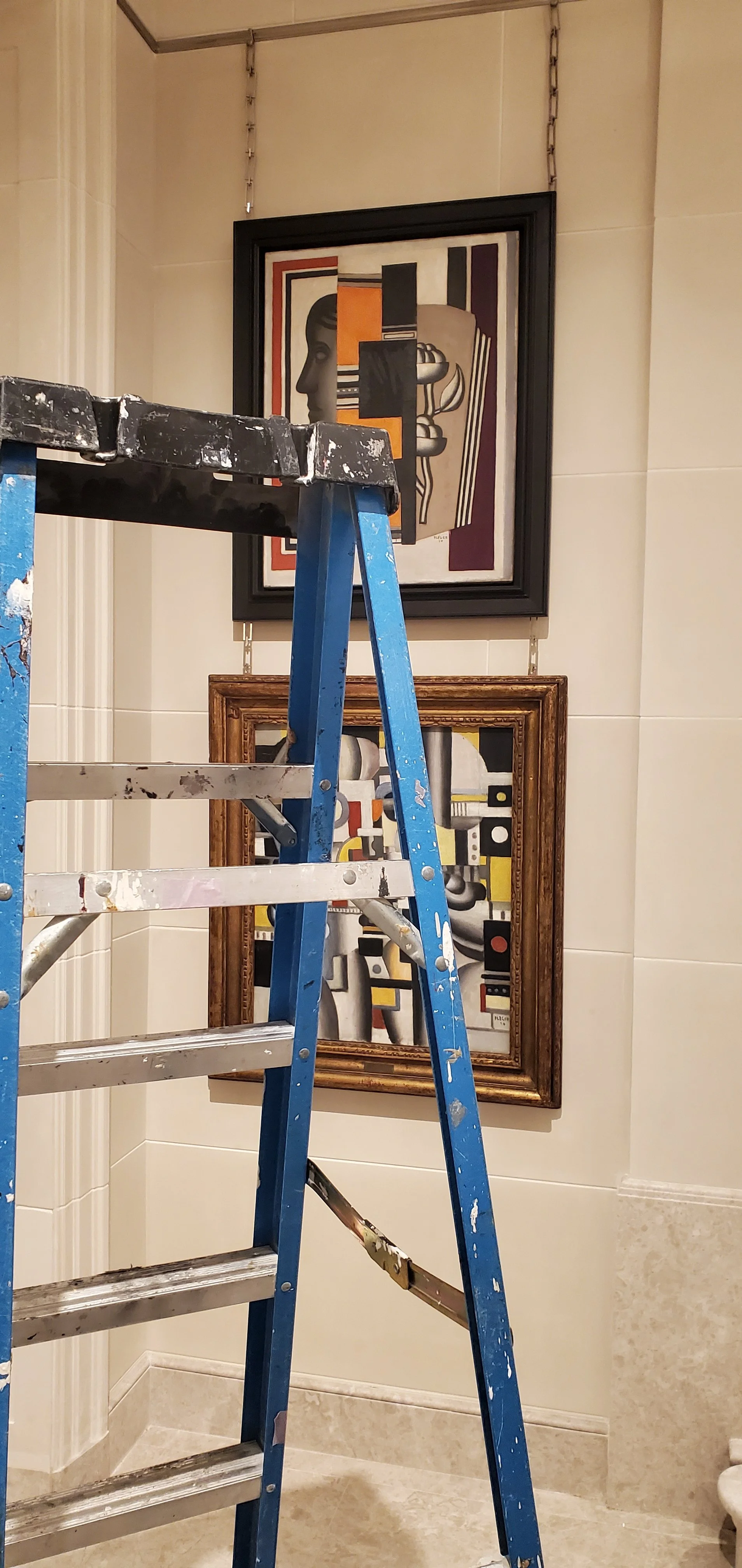 Ladder in front of two framed abstract paintings hanging on a beige tiled wall.