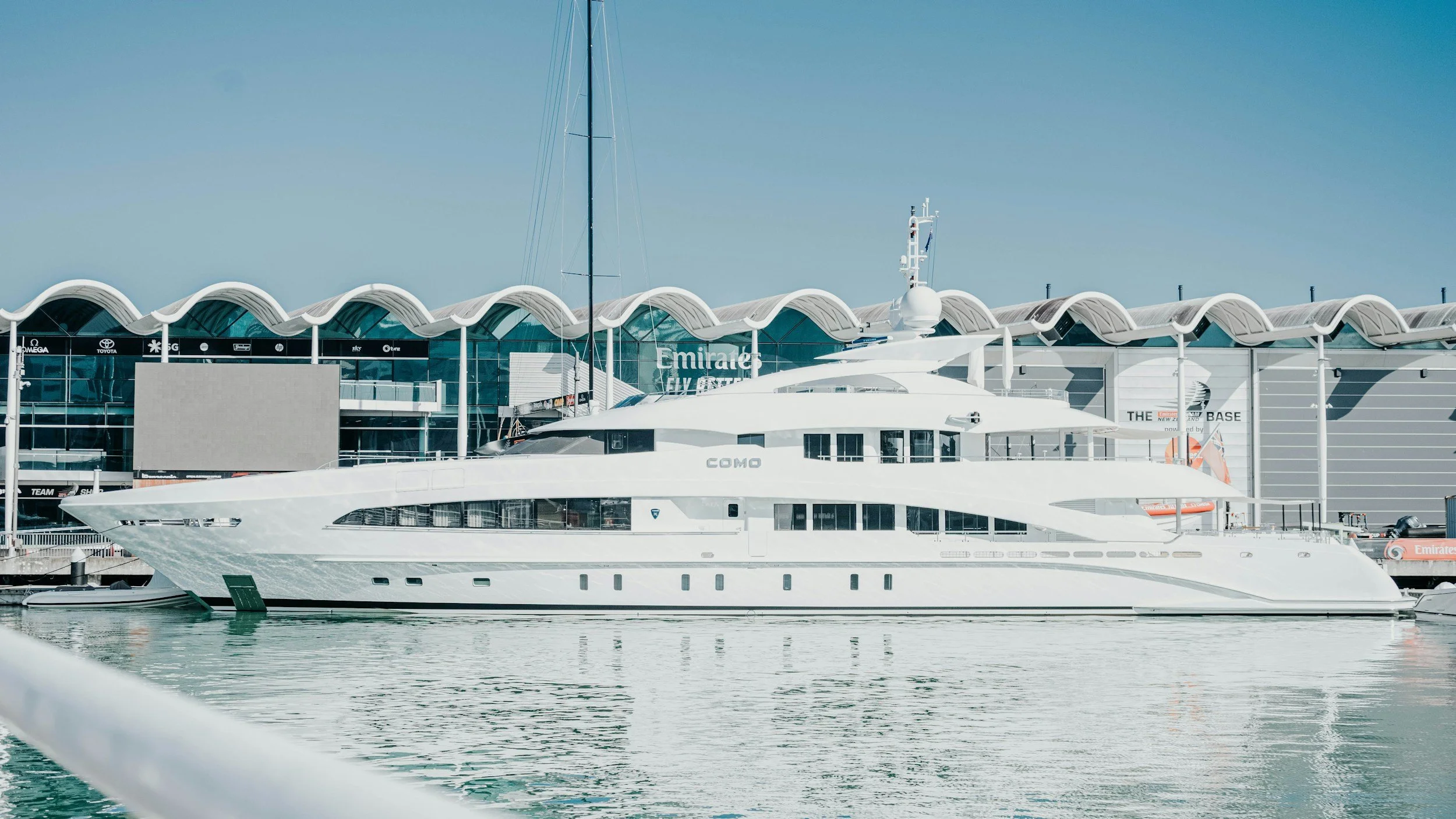 A large white yacht docked at a marina with a modern building in the background, featuring a wave-like roof design and various signs.