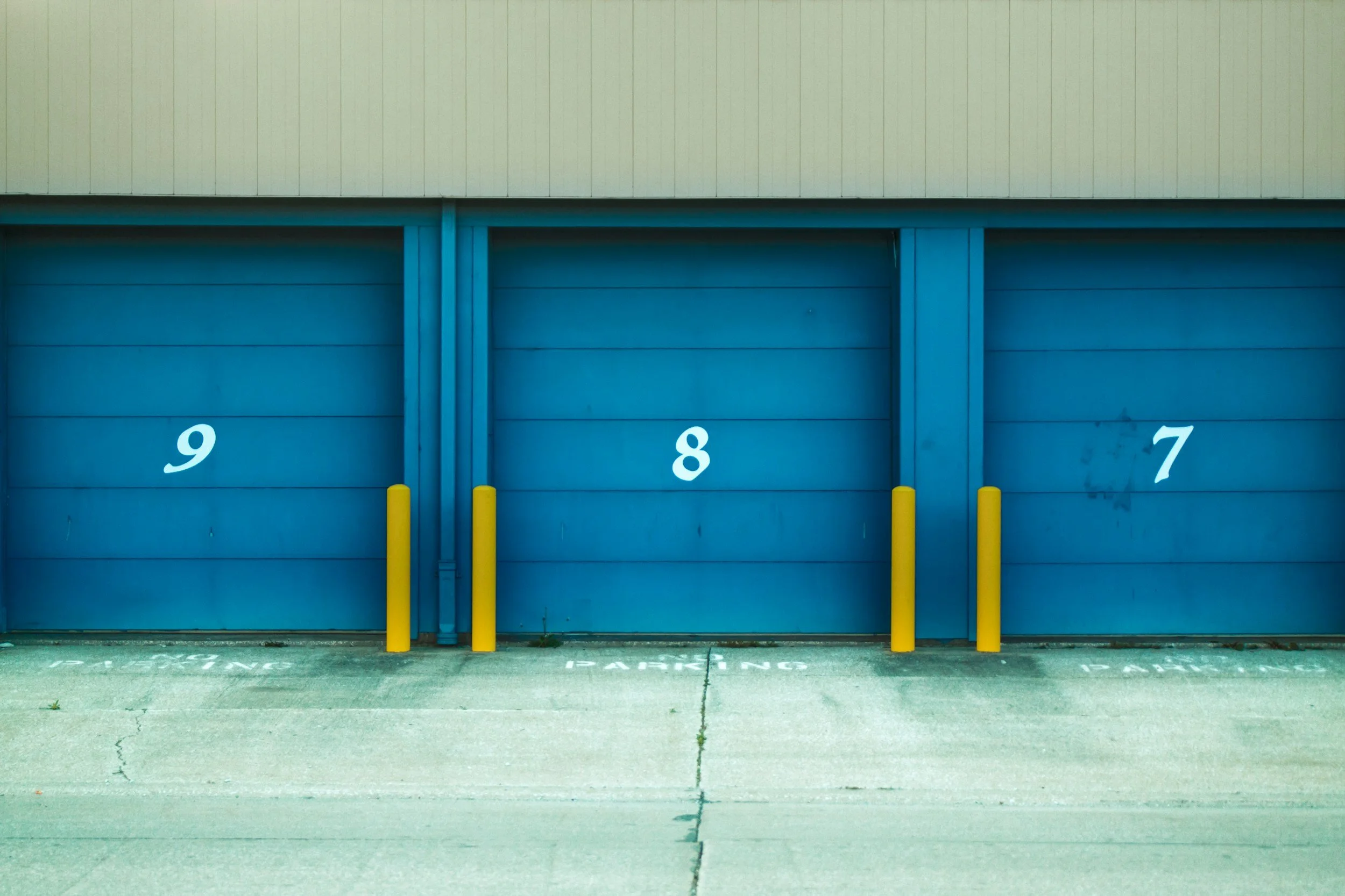 Shelving units with boxed items stored in a warehouse or storage area. The boxes have bright labels, and the background features a bare concrete wall.