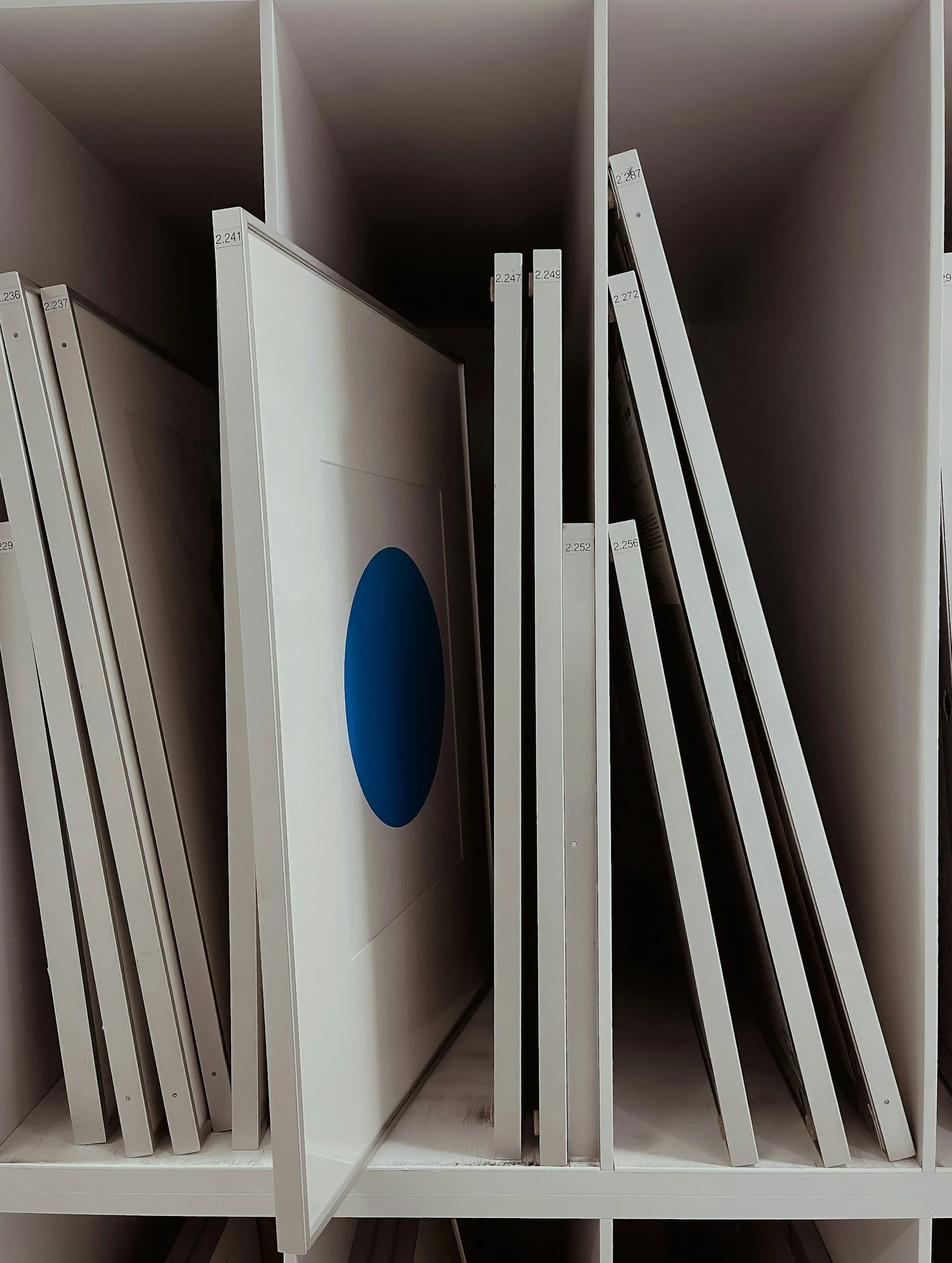 Empty art storage room with white metal racks, paintings, a step ladder, and a rolling cart.