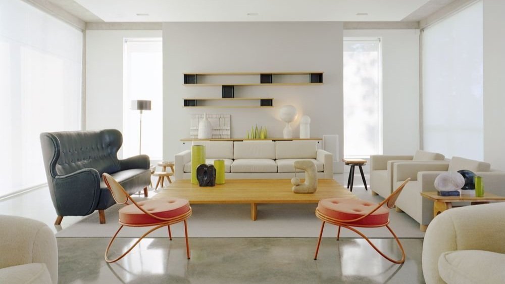 Modern living room with white walls, large windows, a white sofa, a black high-back armchair, two coral-colored chairs, and a wooden coffee table with decorative objects.