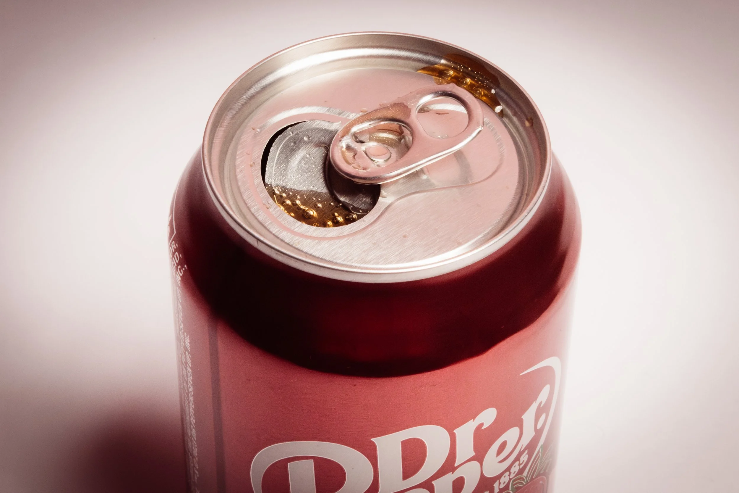 Close-up of the top of a red can of Dr. Pepper soda showing the pull tab open with some soda inside.