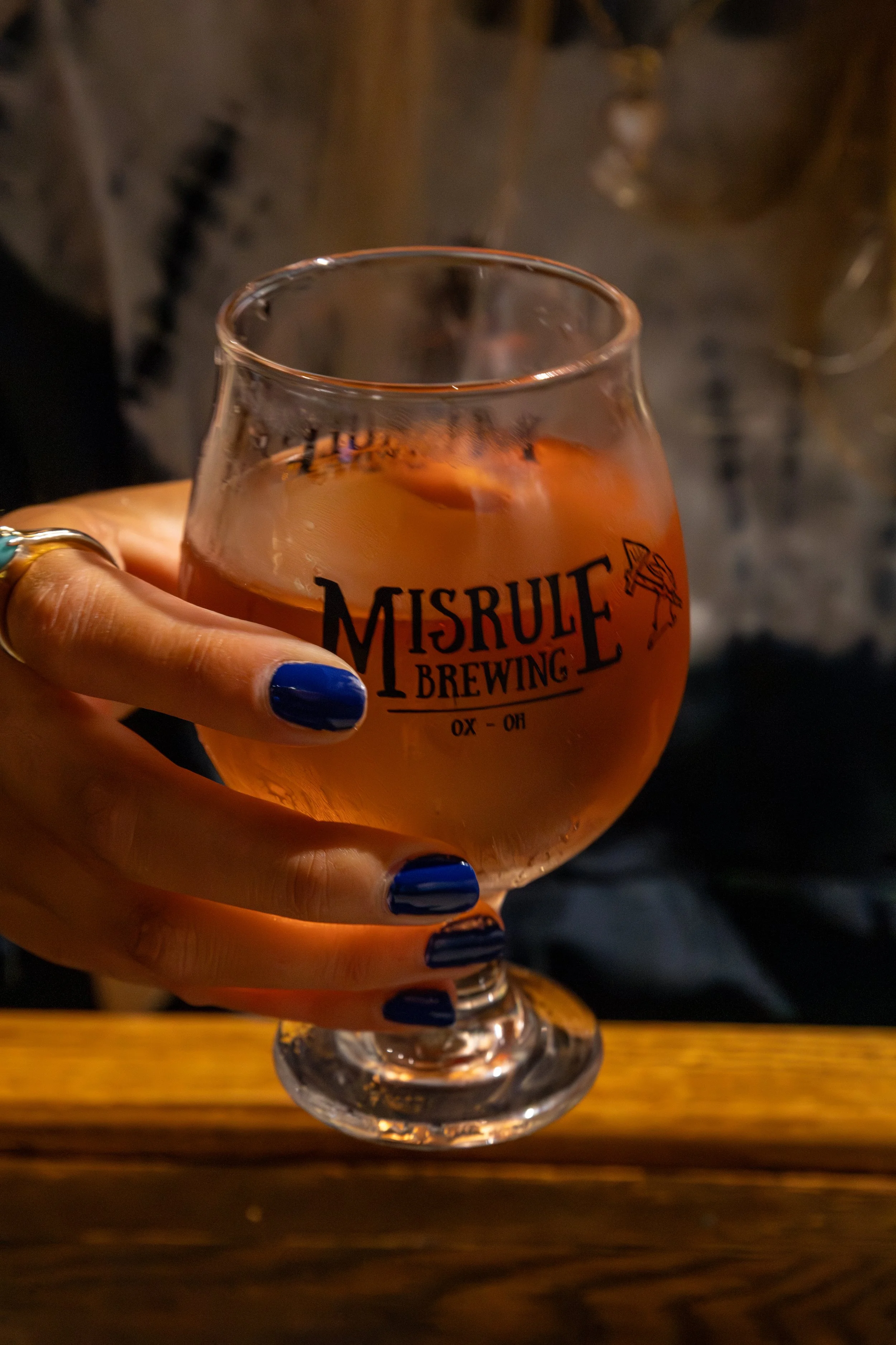 A person with blue painted nails holding a rounded glass of pink cider from Misrule Brewing, with a wooden surface in the foreground and a blurred dark background.