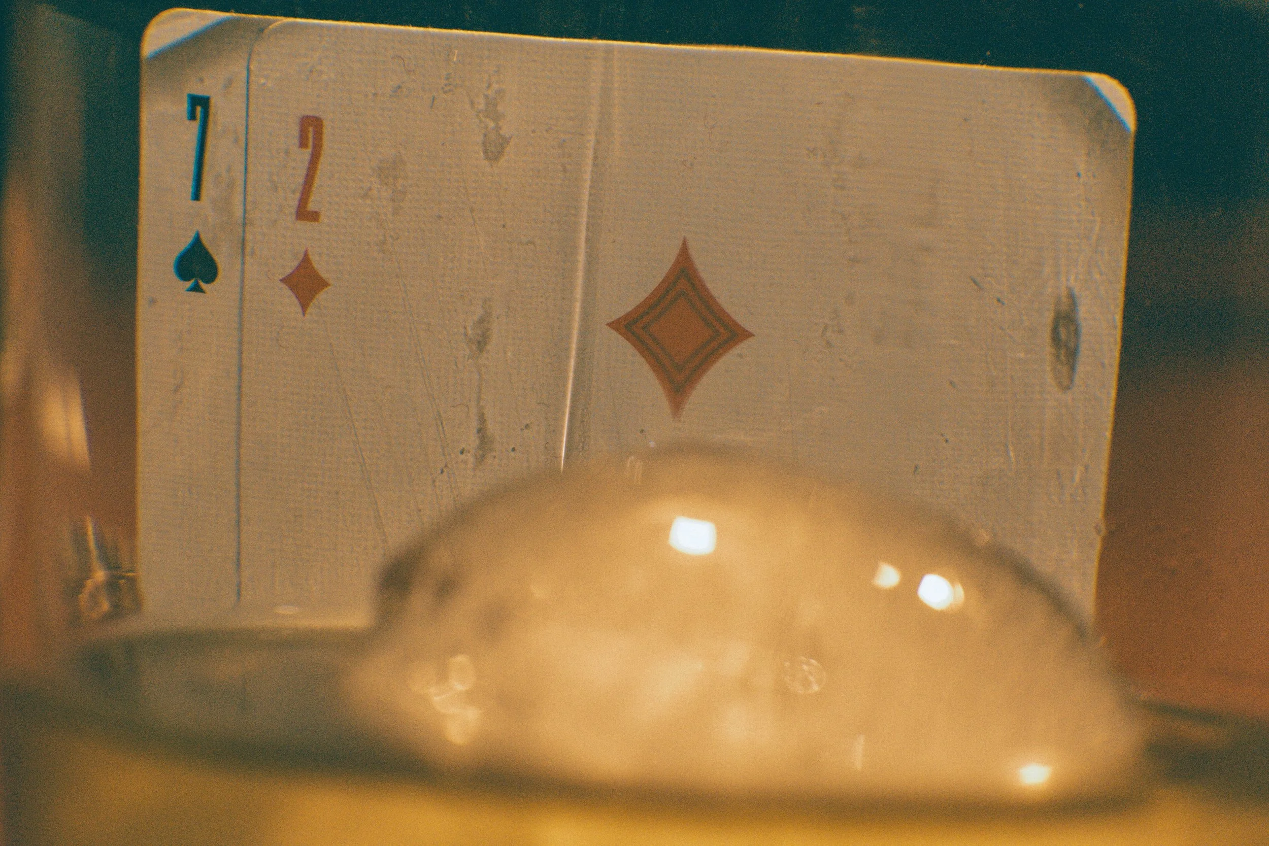 Close-up of a deck of playing cards, showing the Ace of Spades, the 2 of Diamonds, and the 7 of Spades, with a glass of whiskey in the foreground.