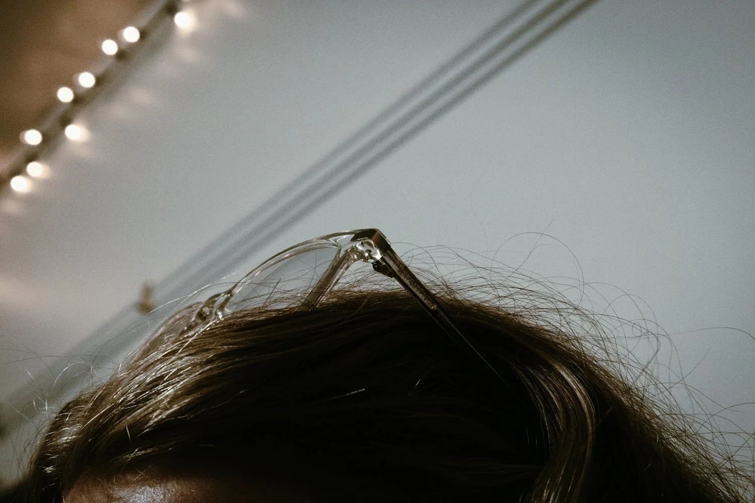 Close-up of a person's head with brown hair and clear glasses resting on top of their head, with string lights visible in the background.