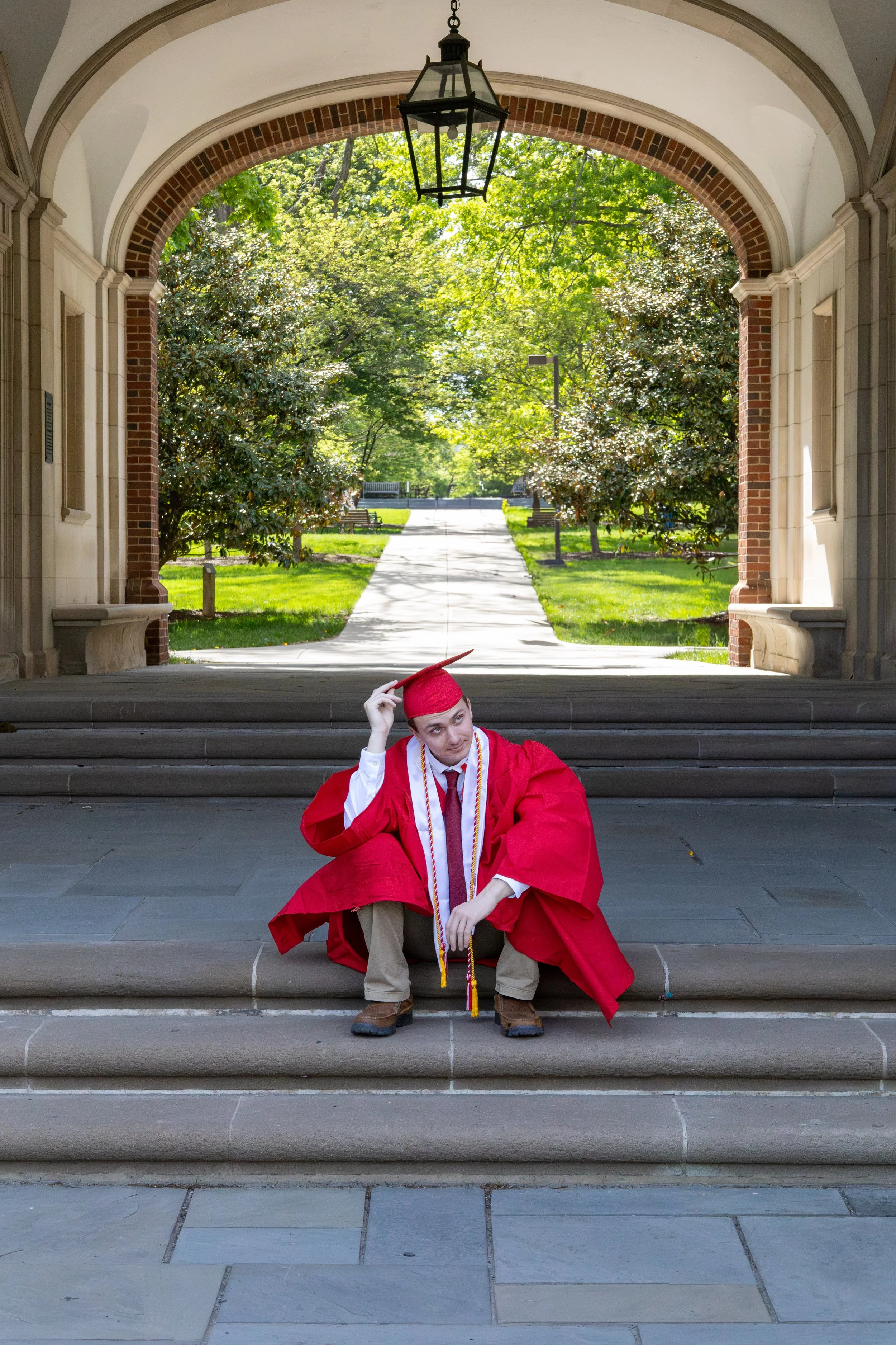 A young man dressed in a red graduation gown and cap, sitting on the steps of a building under an archway, with a pathway and trees in the background.
