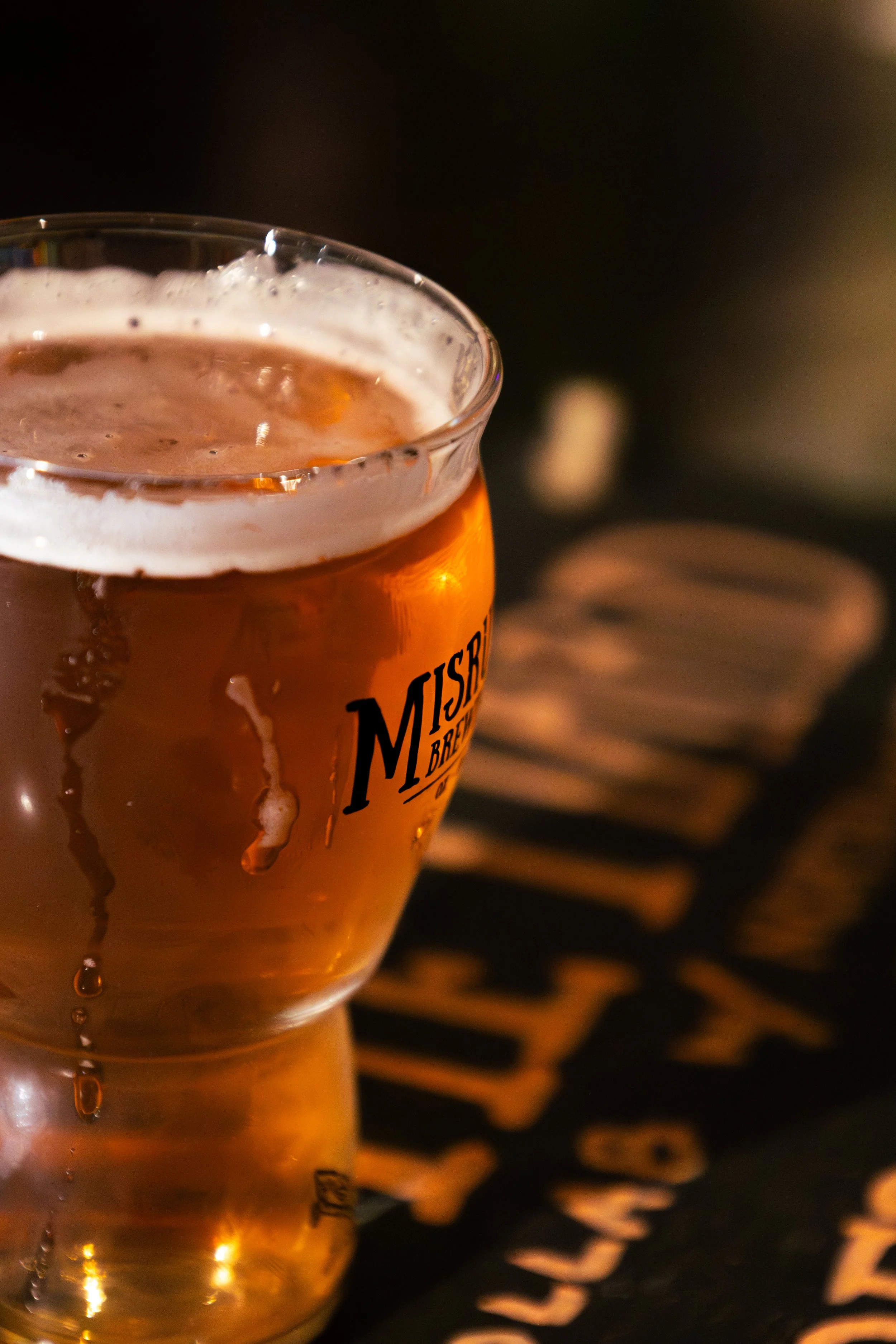 A glass of beer on a dark table with a logo reading 'MisruleBrewing' and a blurred background.