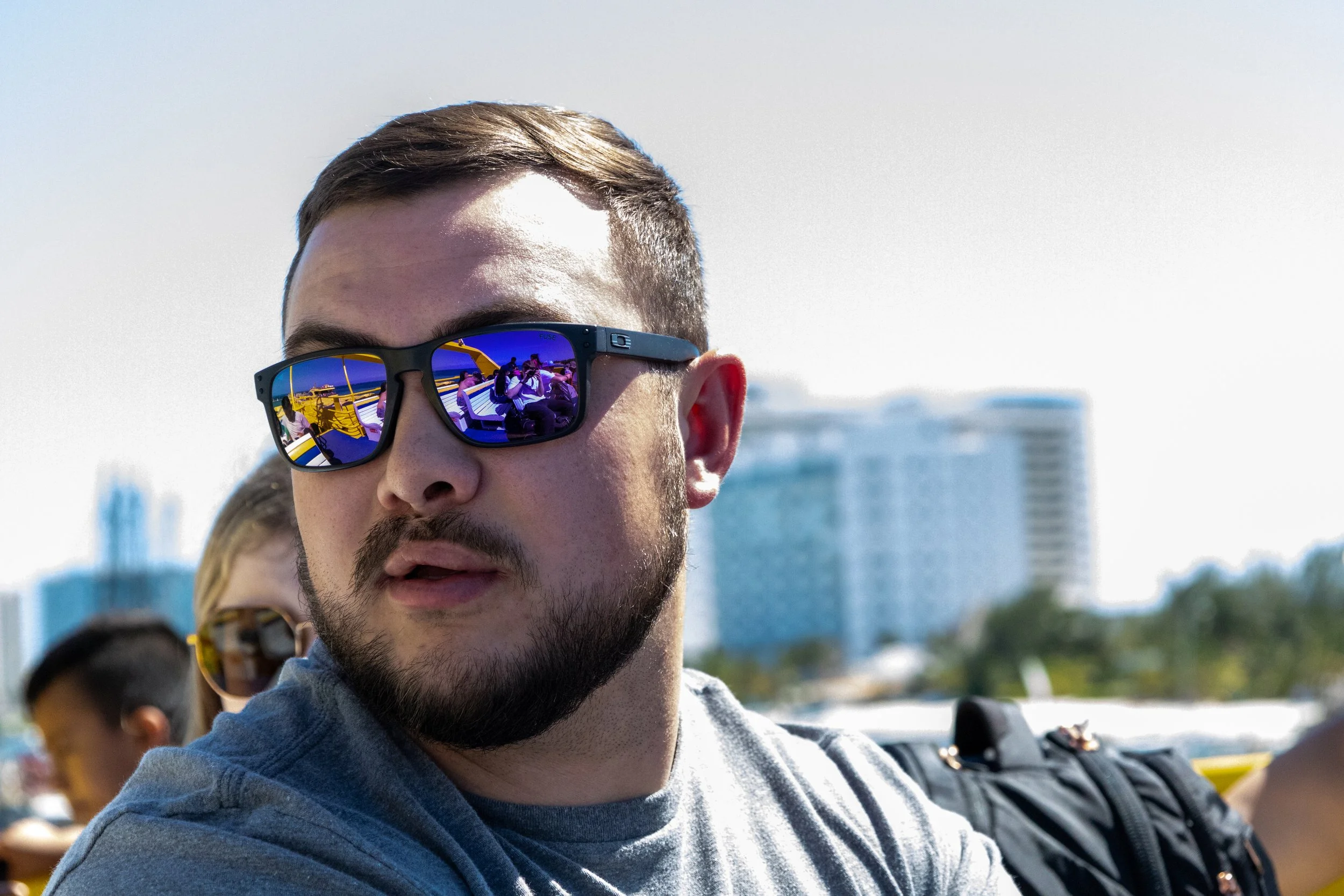 A man with a beard and sunglasses with reflective blue lenses, with a woman with sunglasses behind him, on a boat or outdoor area, with city buildings in the background.
