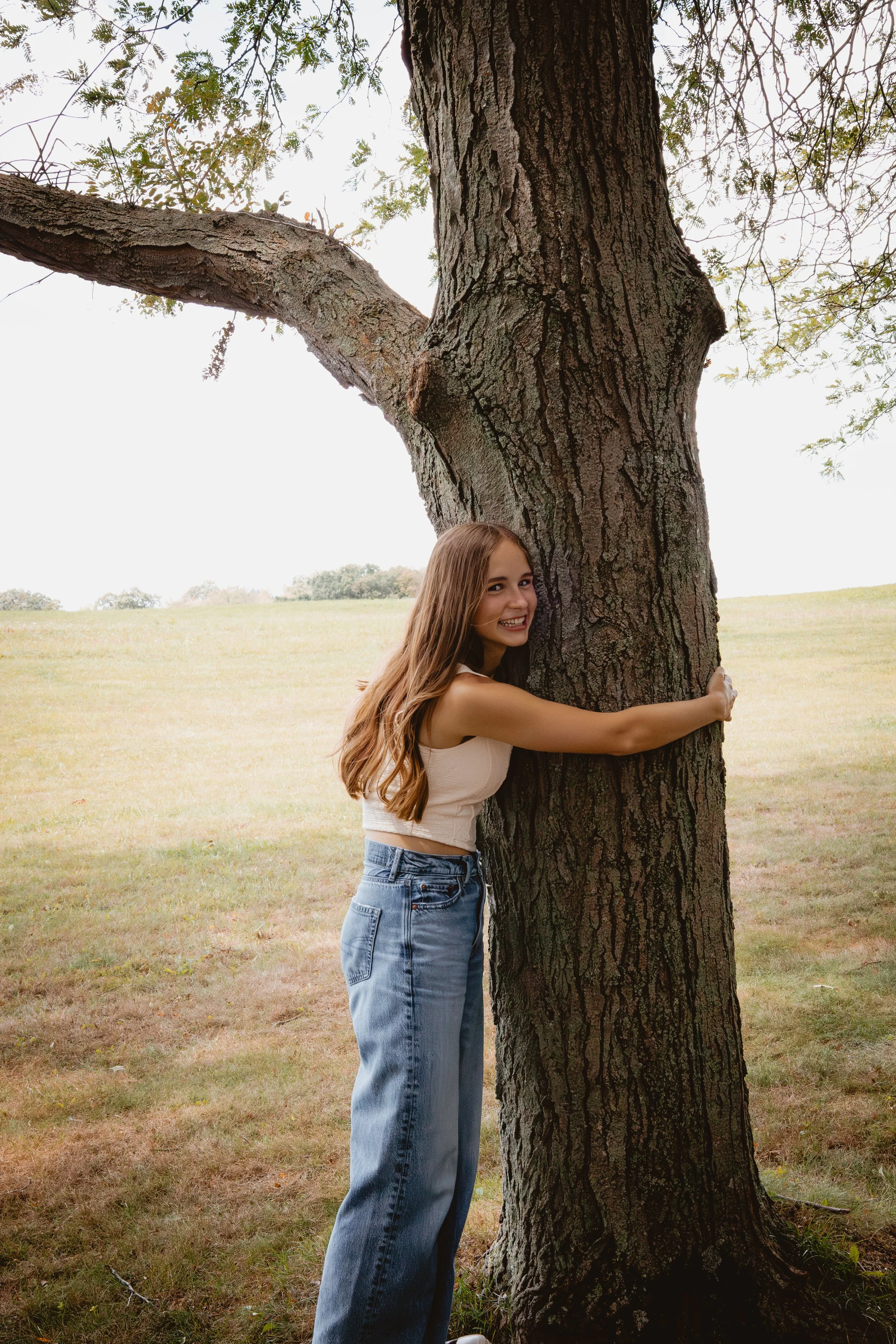 A smiling young woman hugging a large tree trunk in an open grassy field.