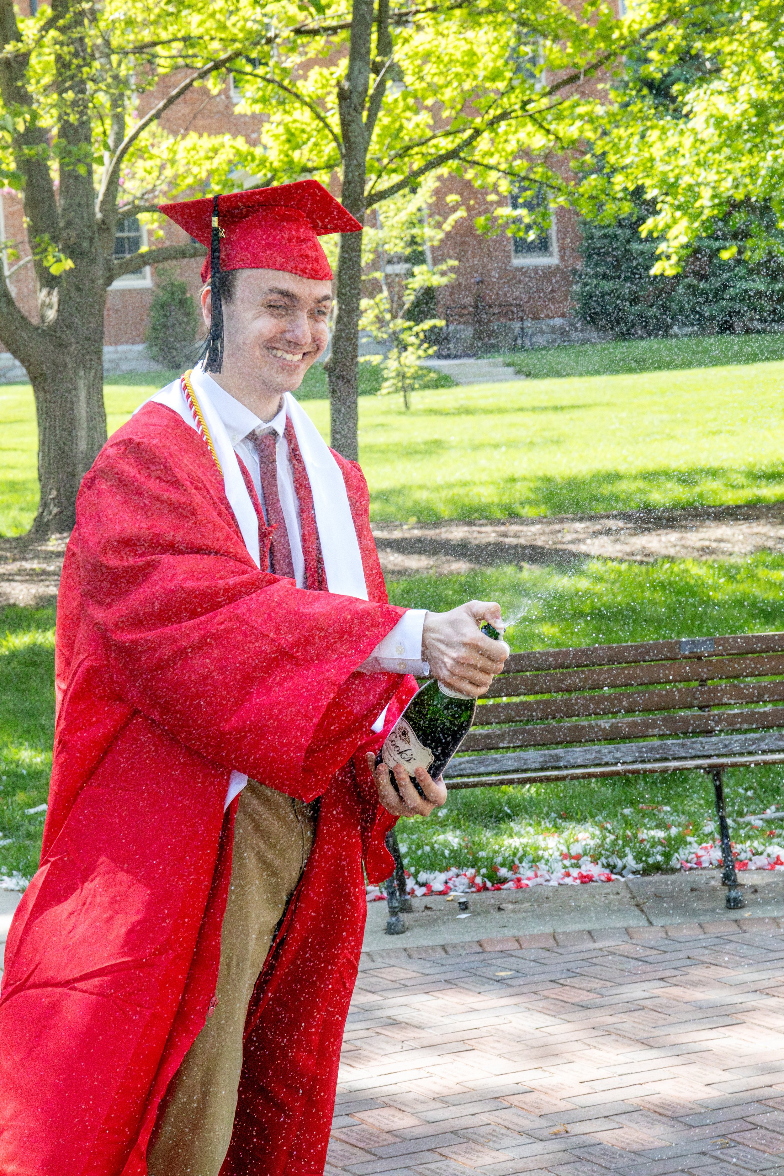 A man in a red graduation gown and cap celebrating outdoors with a bottle of champagne, spraying it as he smiles.
