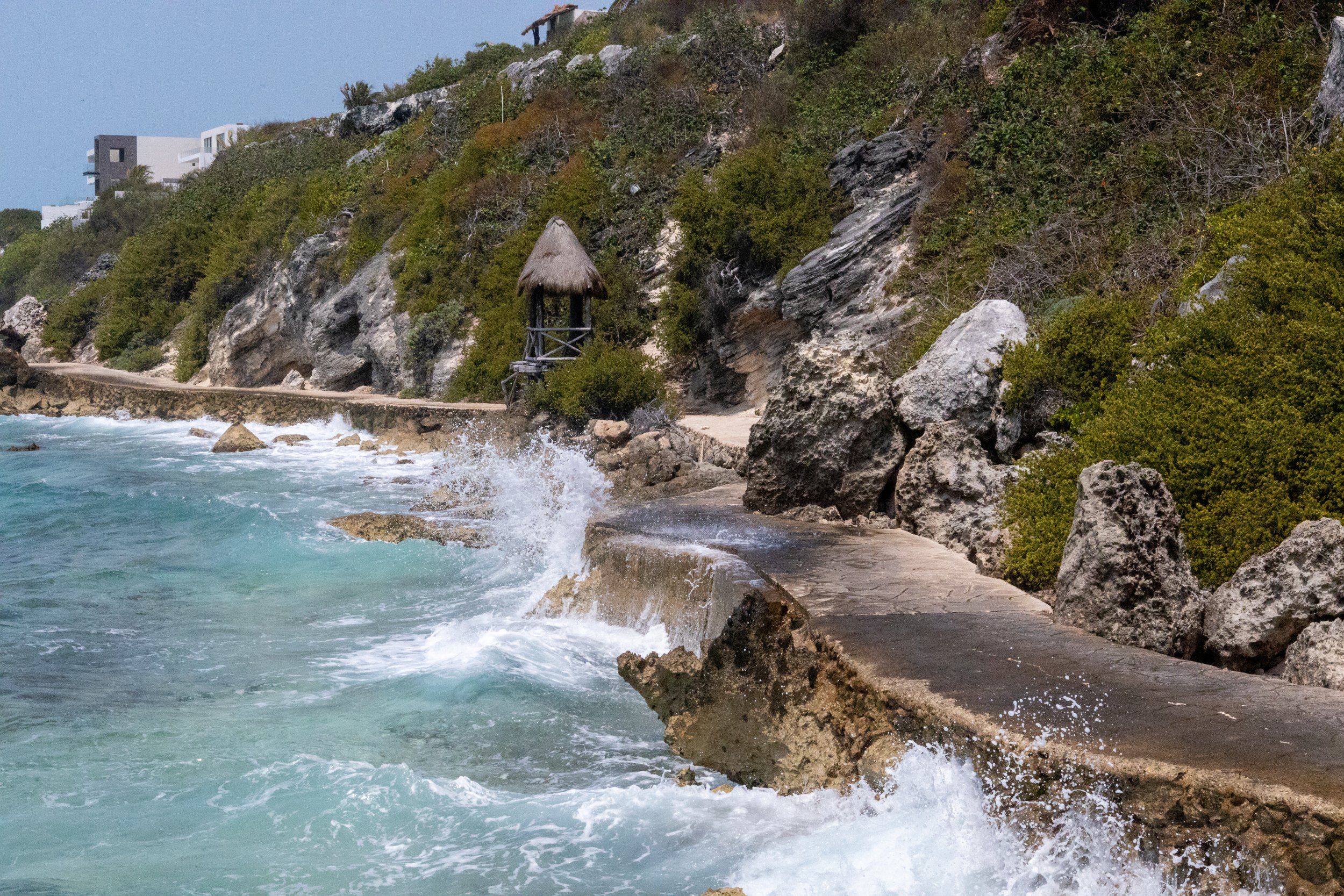 A rocky coastal path beside the ocean with waves crashing against it, leading to a small thatched hut on the hillside surrounded by greenery.