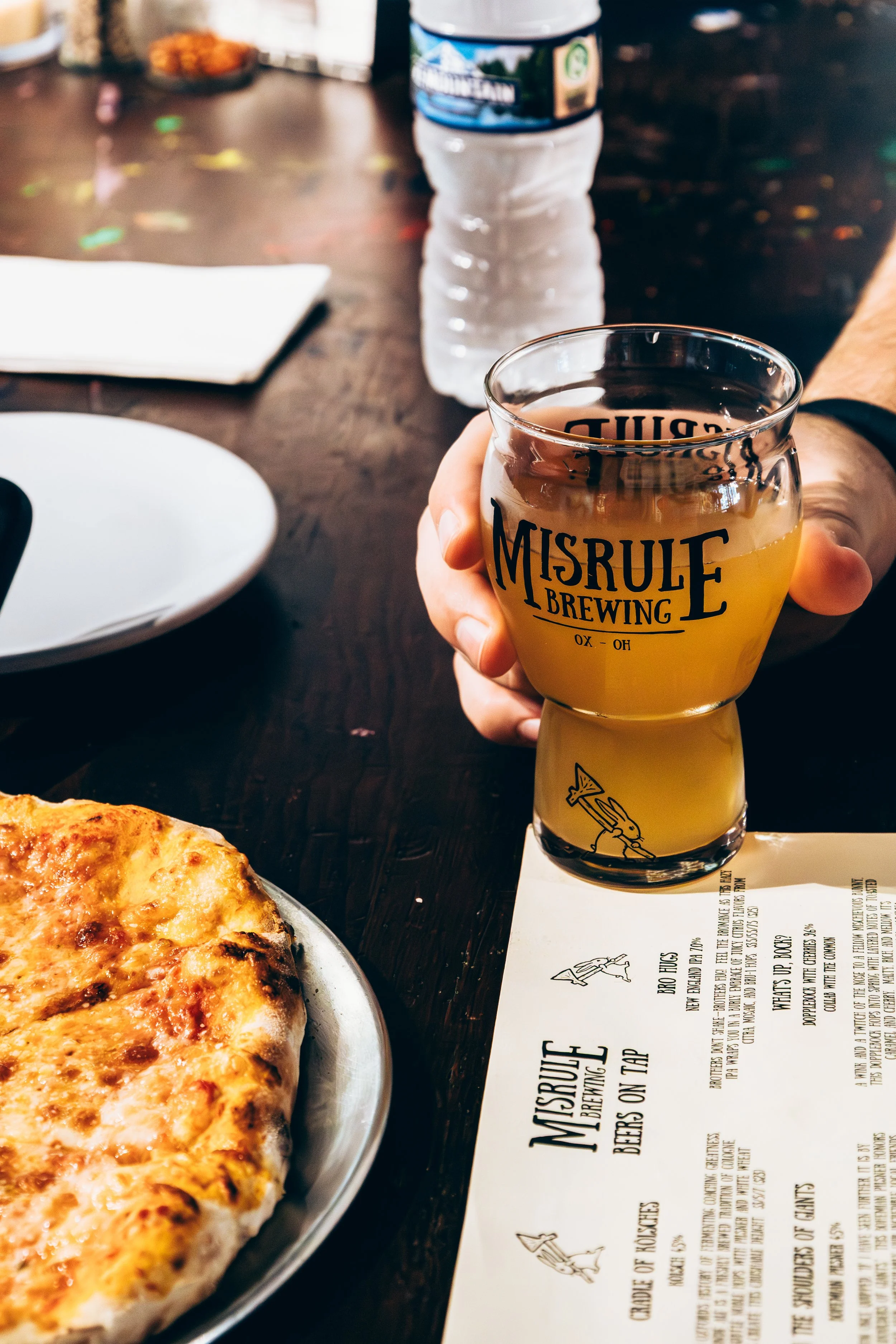 Person holding a glass of beer from Misrule Brewing, with a pizza, a bottle of water, and a menu on the table.