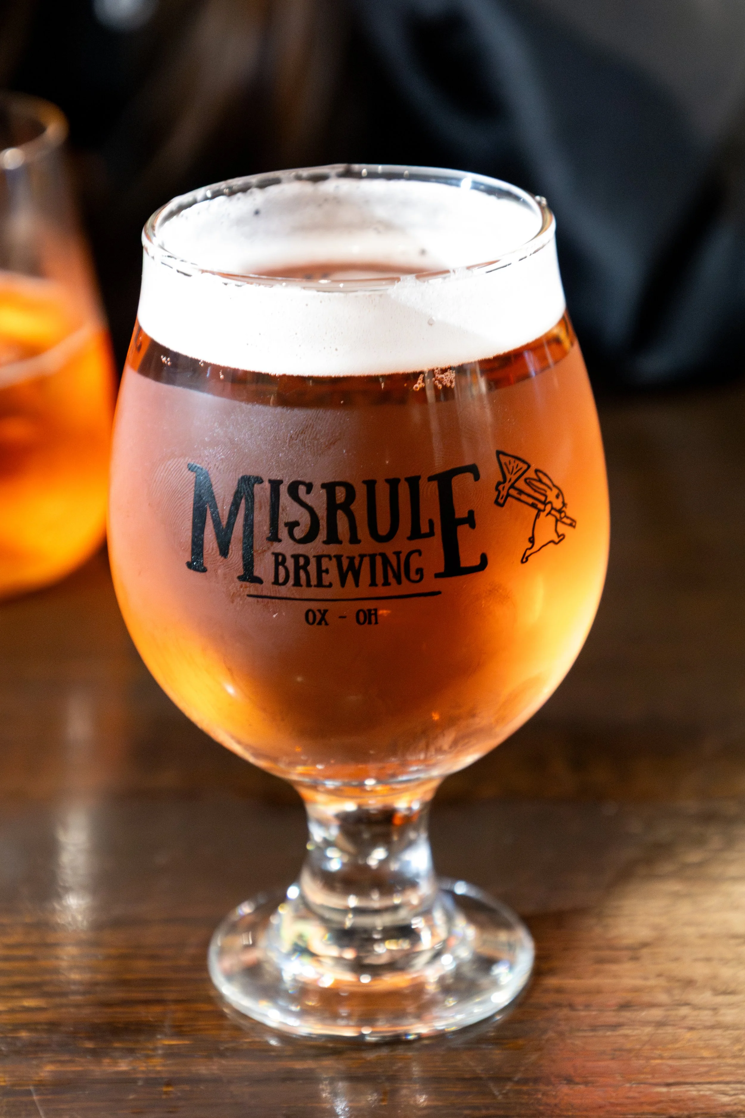 A glass of cider with a label from Misrule Brewing, sitting on a wooden table.