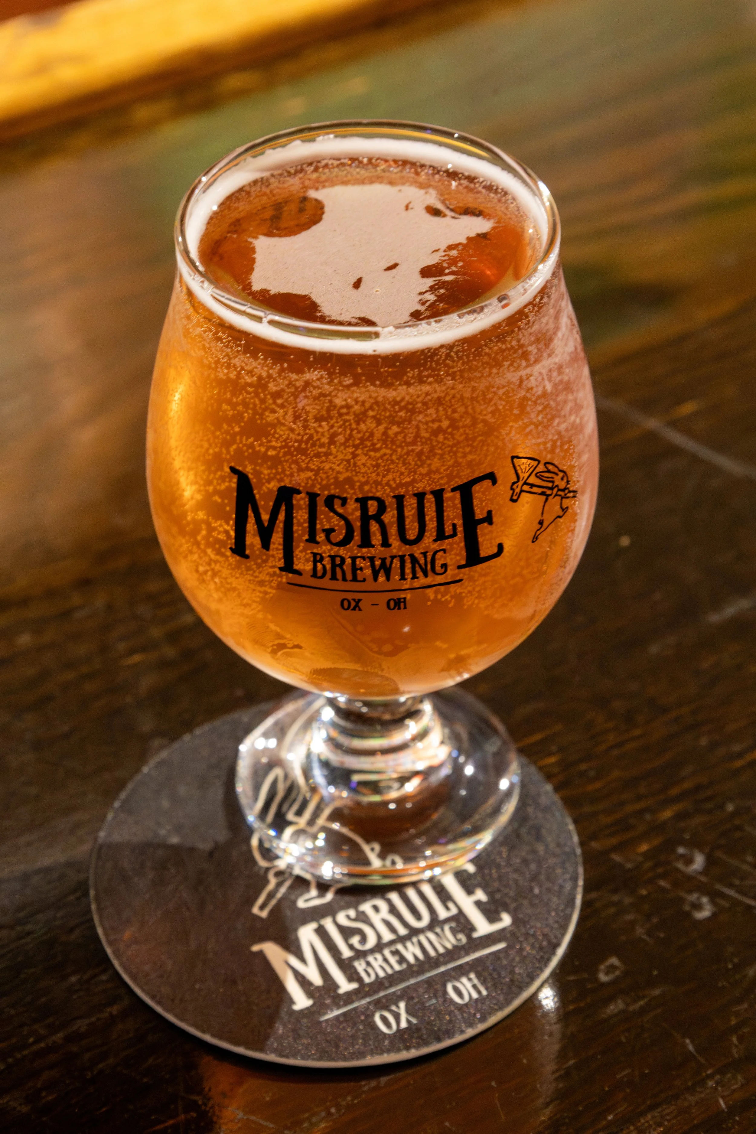 A glass of cider with foam on top, sitting on a coaster on a wooden surface. The glass has the logo 'Misrule Brewing Co.' on it.