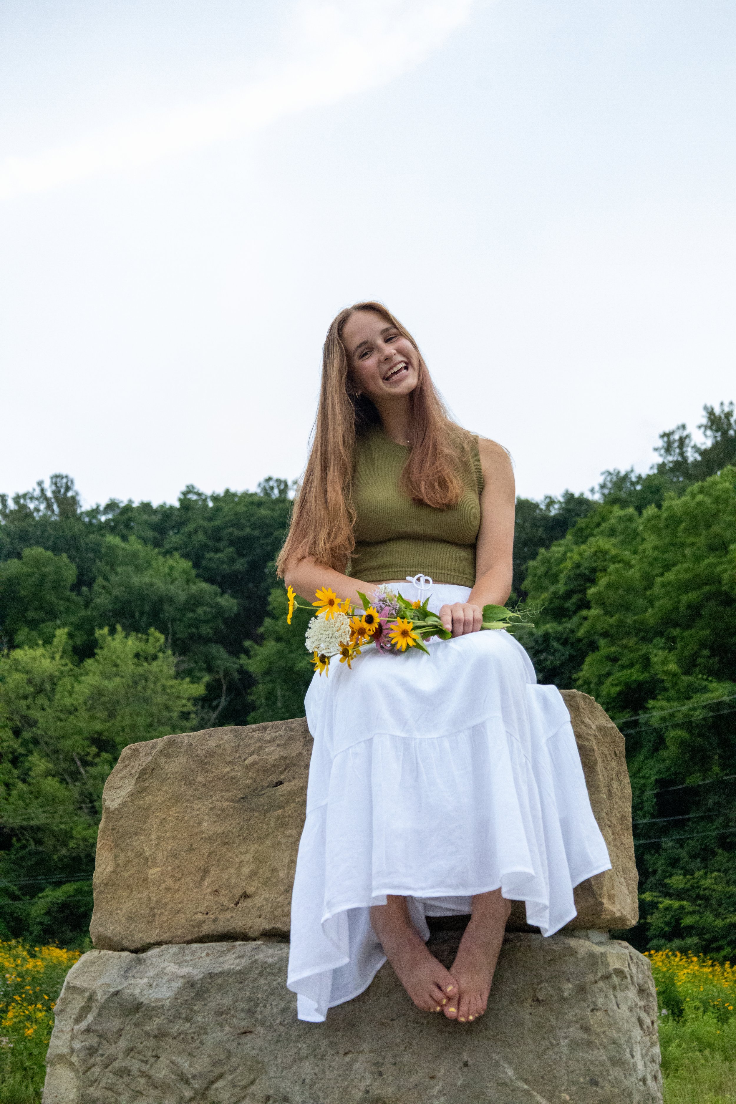 A young woman with long brown hair, wearing a green sleeveless top and a white skirt, sitting on a large rock outdoors surrounded by greenery, holding a bouquet of yellow and white flowers, smiling and looking at the camera.