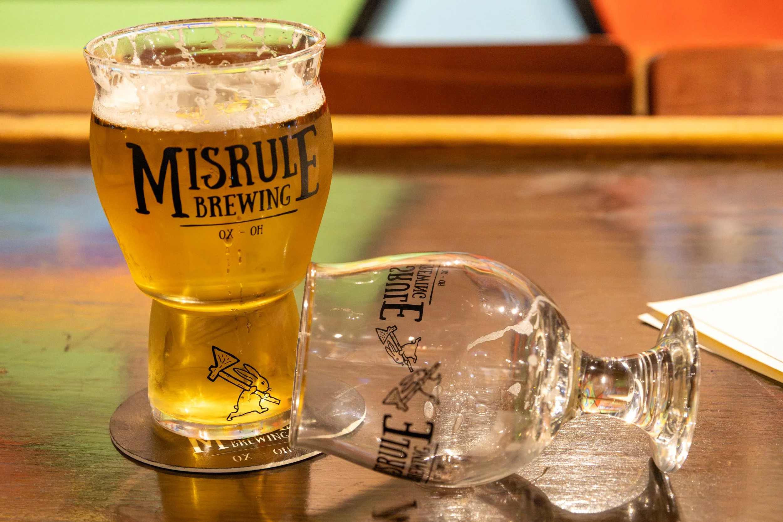 Two glasses, one upright filled with beer and one overturned on a wooden table, both glasses have the logo of Misrule Brewing.
