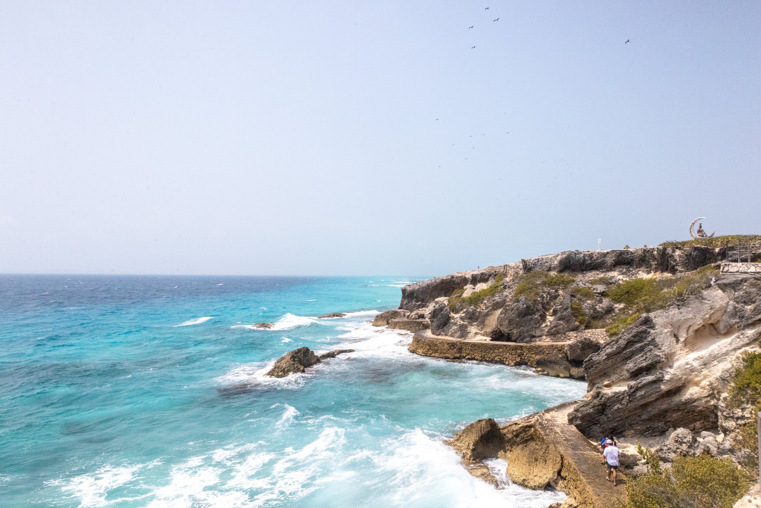 Coastal cliffs with turquoise water crashing against rocks, two people walking on a path near the shore, and a person relaxing on a crescent-shaped sculpture on the cliff top.