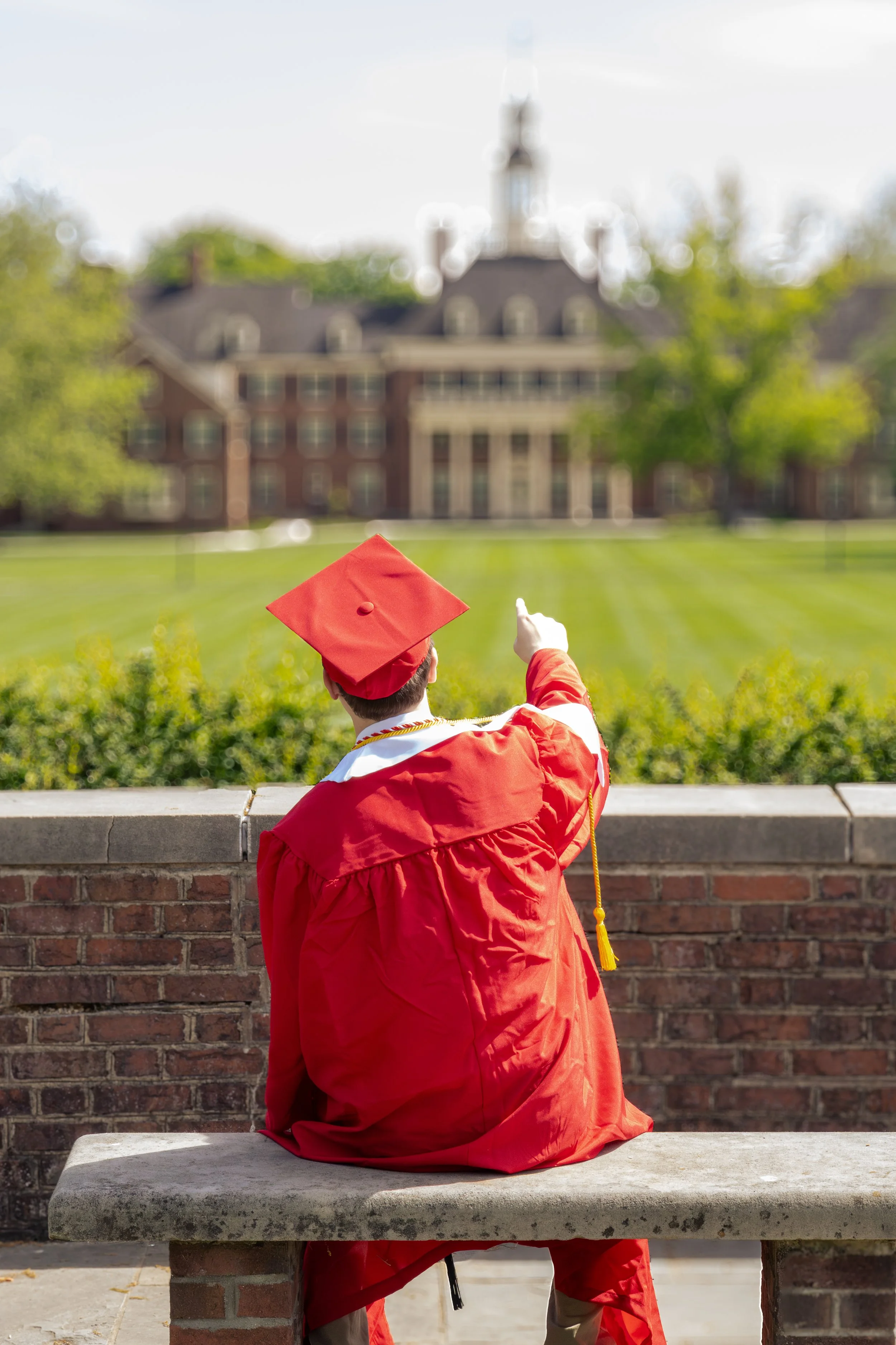 A person in a red graduation gown and cap sitting on a stone bench, pointing towards a large building in the distance with a green lawn in front.