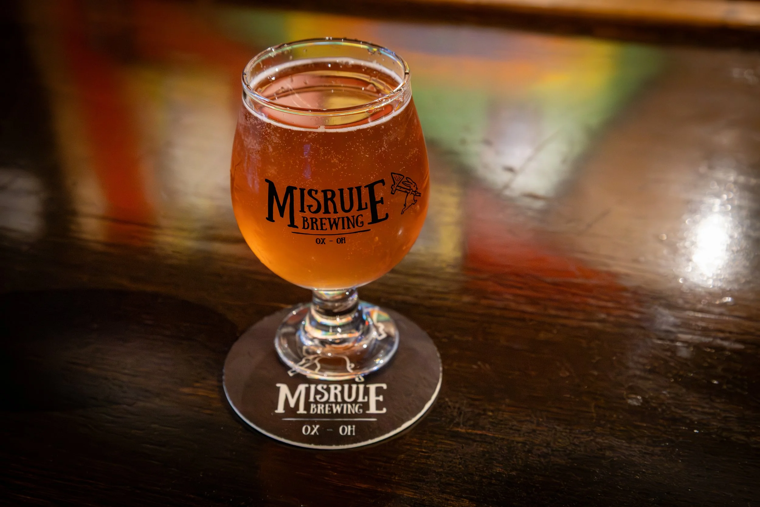 A glass of amber cider on a wooden table with the logo "Misrule Brewing" and the text "OX - OH" on the glass and coaster.