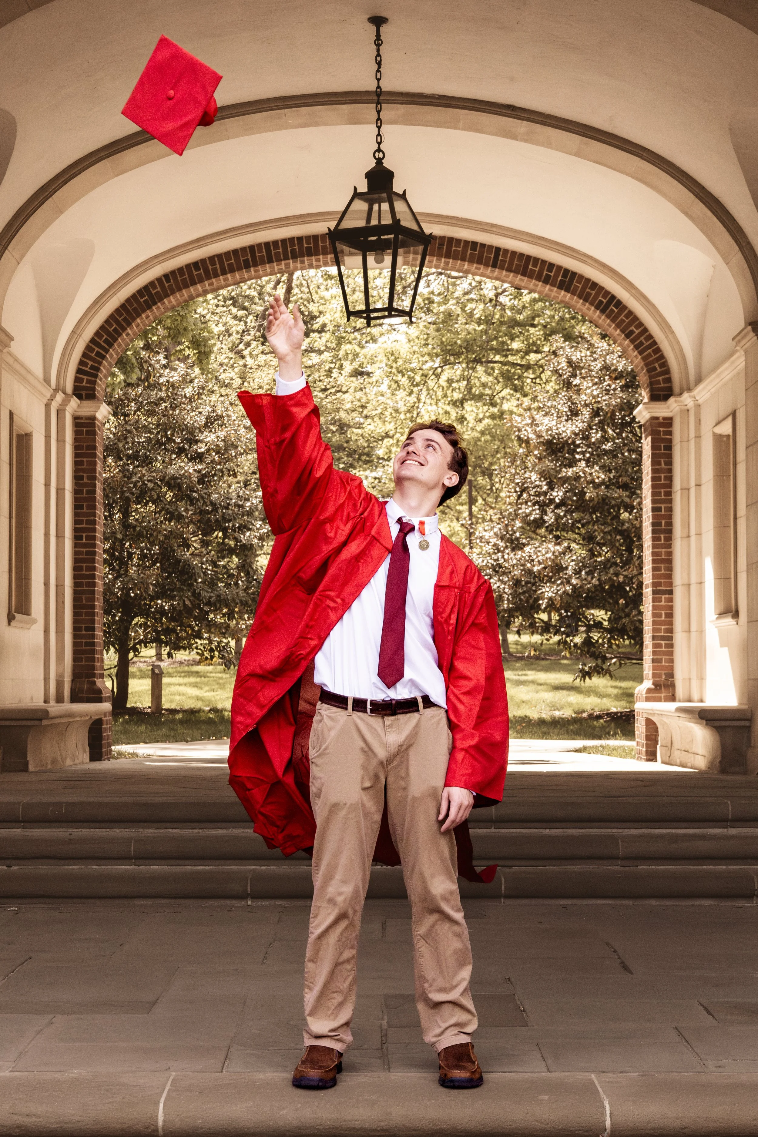 A young man in a red graduation gown and cap throws his cap into the air and smiles under an arched walkway with trees in the background.