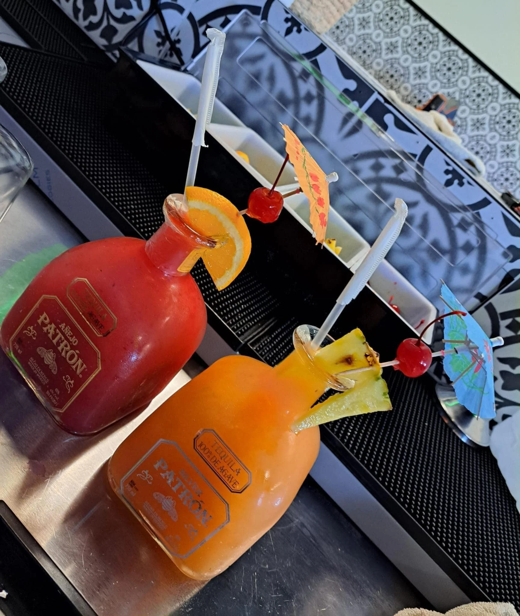 Two large glass bottles of tropical fruit cocktails with garnishes, mini umbrellas, cherries, pineapple, and orange slices, on a table with a tray of condiments in the background.