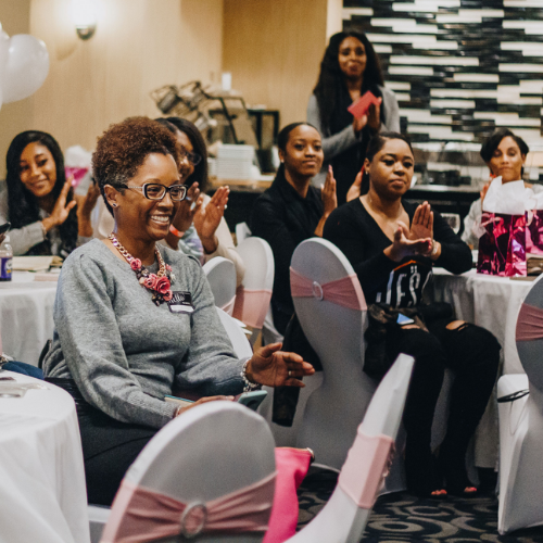 Group of women seated at tables in a conference room, clapping and smiling, with a woman standing in the background holding a pink folder.
