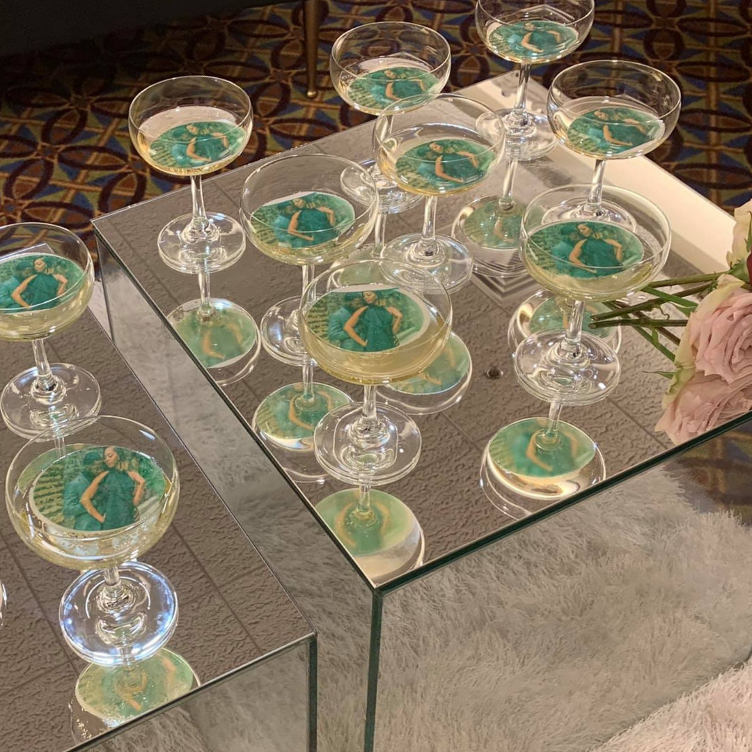 Several champagne glasses with photos of a woman in a green dress floating on top of the drink, arranged on a glass table with a mirror surface.