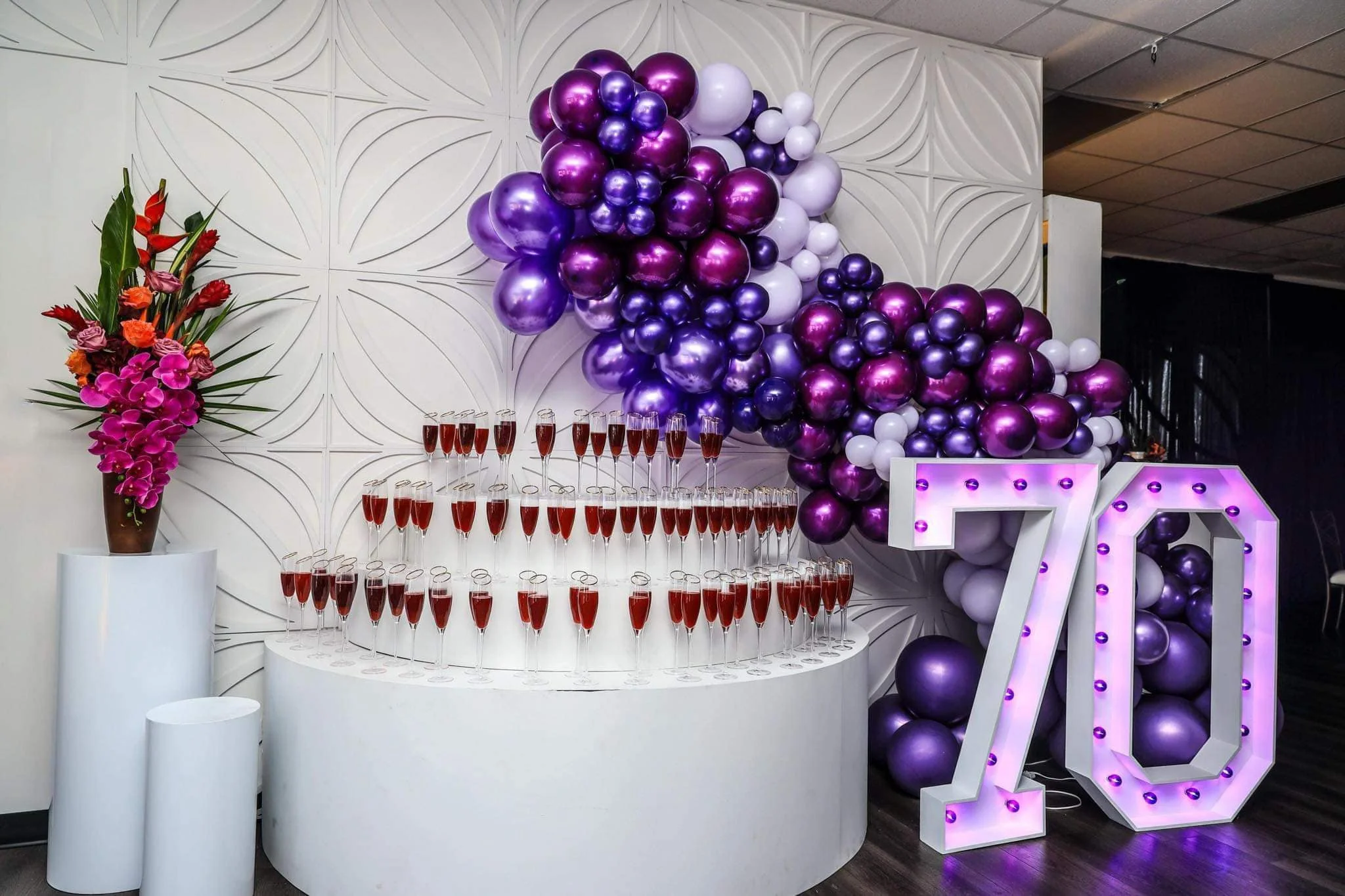 Decorative backdrop with a balloon arrangement in purple, white, and pink, a large illuminated '70' sign, a flower arrangement with red and purple flowers in a tall brown vase, and a tiered display of champagne flutes filled with pink beverage for a 70th birthday celebration.