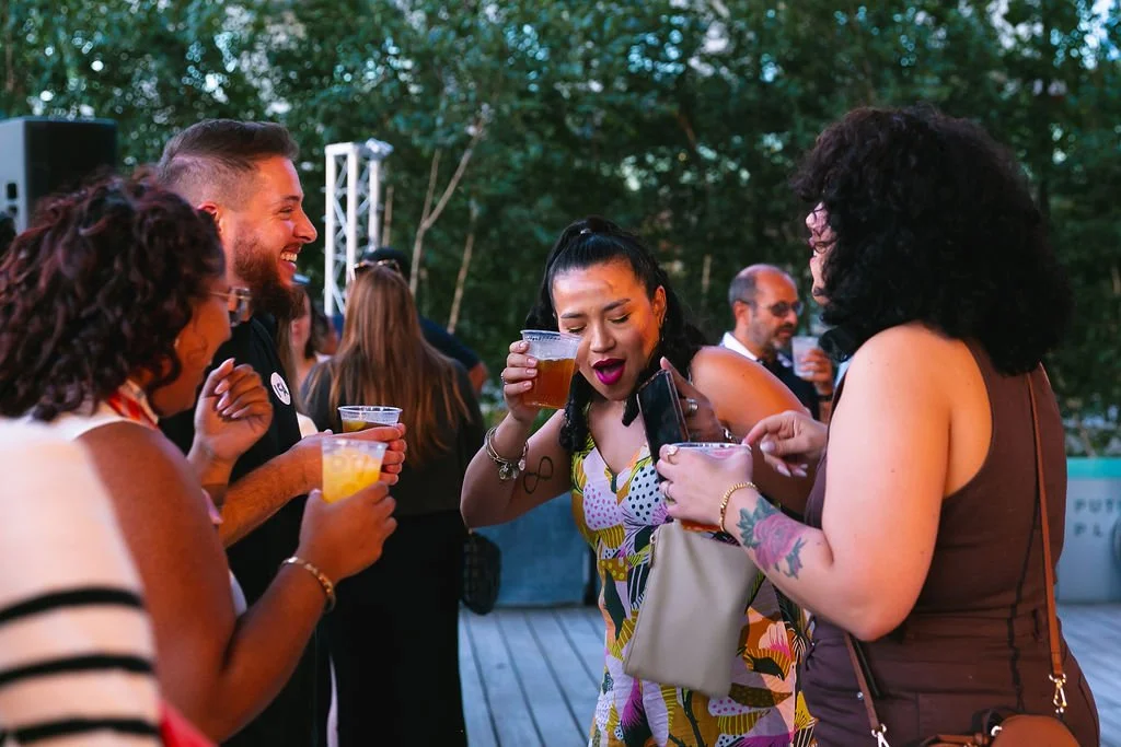 Four people are at an outdoor party, holding drinks and socializing, with trees in the background.