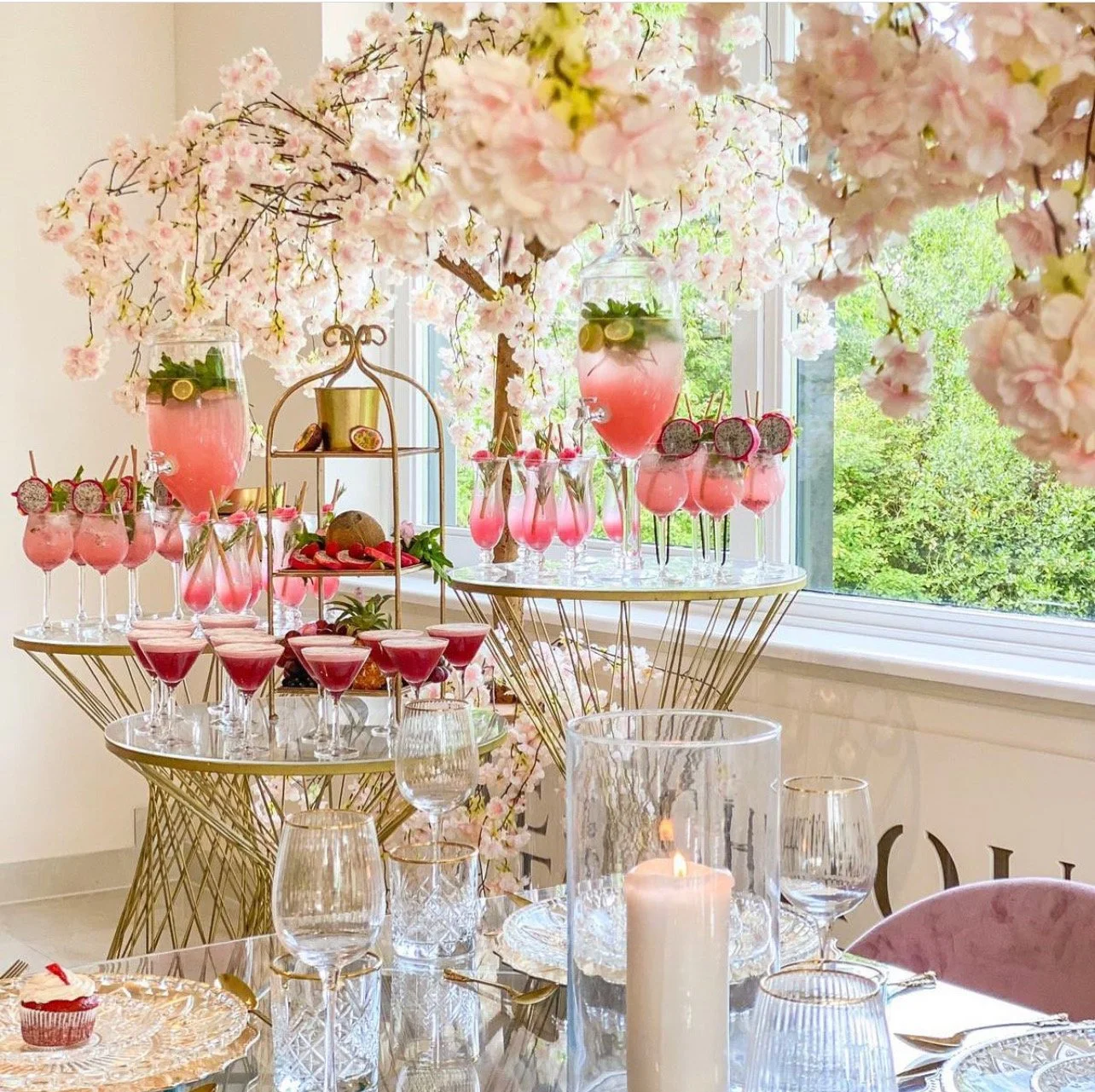A bridal shower or party table decorated with a pink cherry blossom theme, featuring an abundance of pink drinks on gold wire tables, floral centerpieces, and a lit candle on the table.