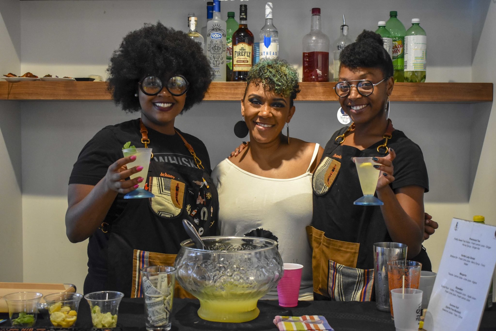 Three women smiling at a bar or beverage station, holding cocktails, with bottles of alcohol and mixers on shelves behind them, suggests a social gathering or party.
