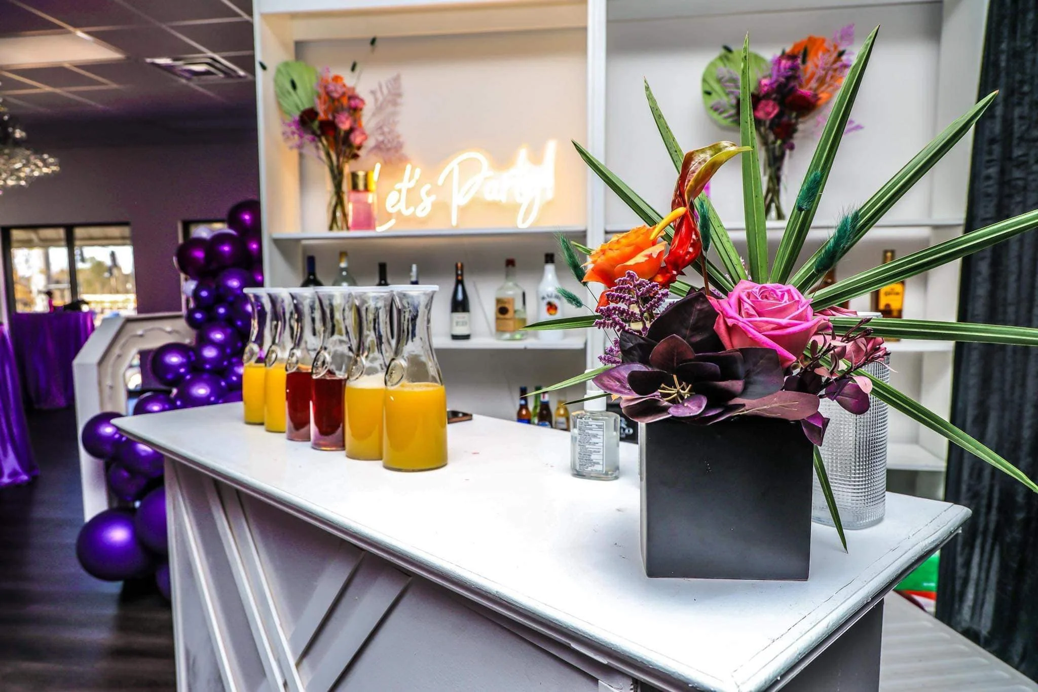 A white bar counter with a black flower arrangement in a black vase, containing pink roses, orange calla lilies, and greenery. Behind the counter are bottles of alcohol, a neon sign that says 'It's Party,' and purple balloons. In the background, there is a window and some purple tablecloths.