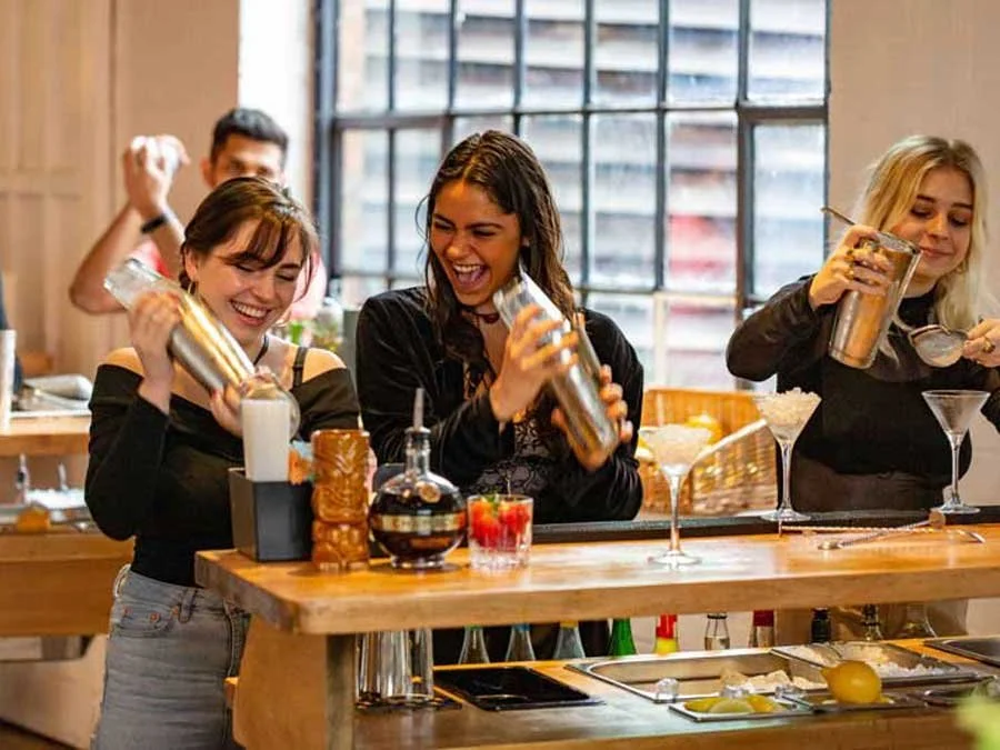 Three women and one man behind a bar counter, smiling and preparing cocktails, with ingredients and glasses on the counter, in a bright, commercial setting.