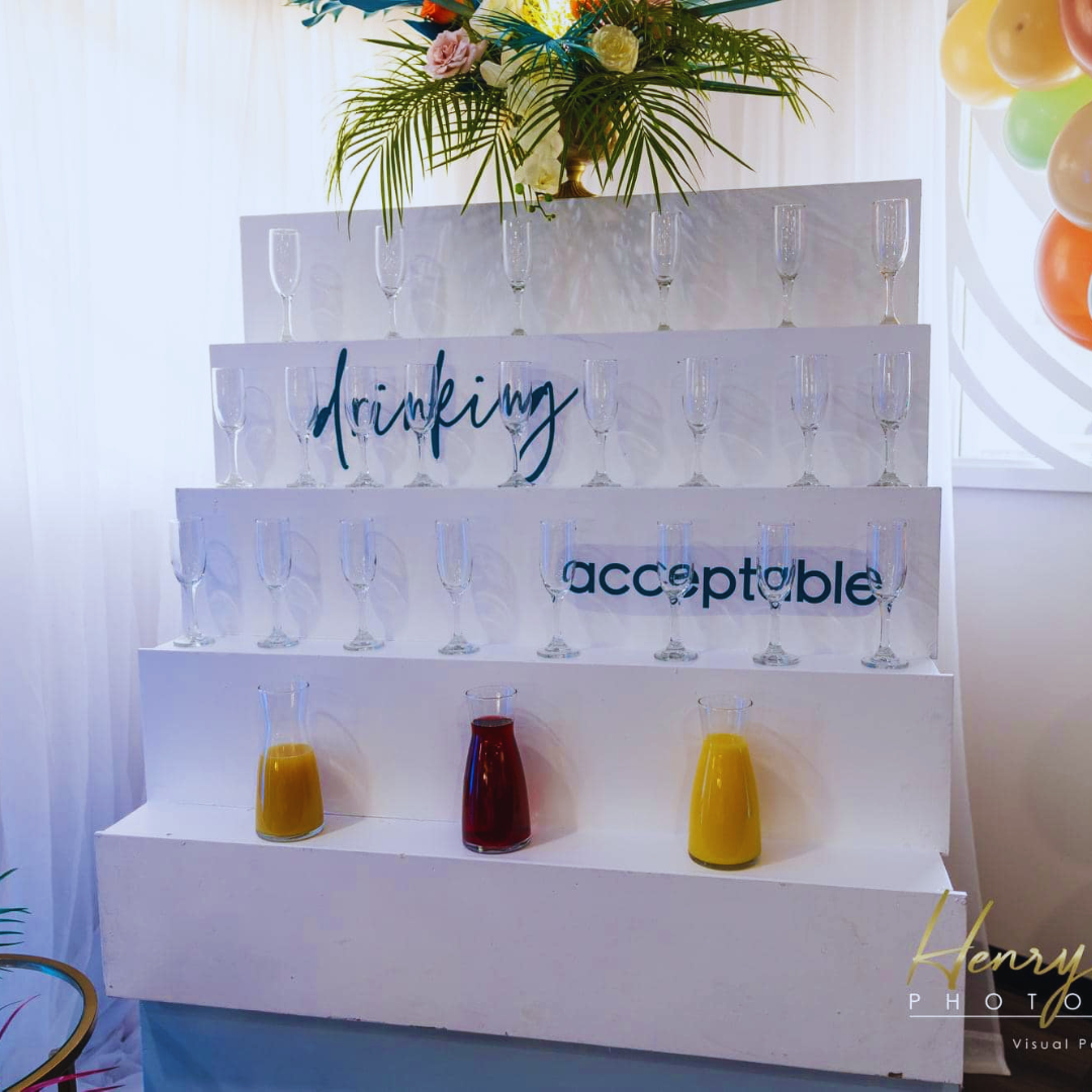 Table with empty champagne glasses on a tiered display with the words 'drinking acceptable' and three pitchers of juice on the bottom tier, decorated with a floral arrangement on top.