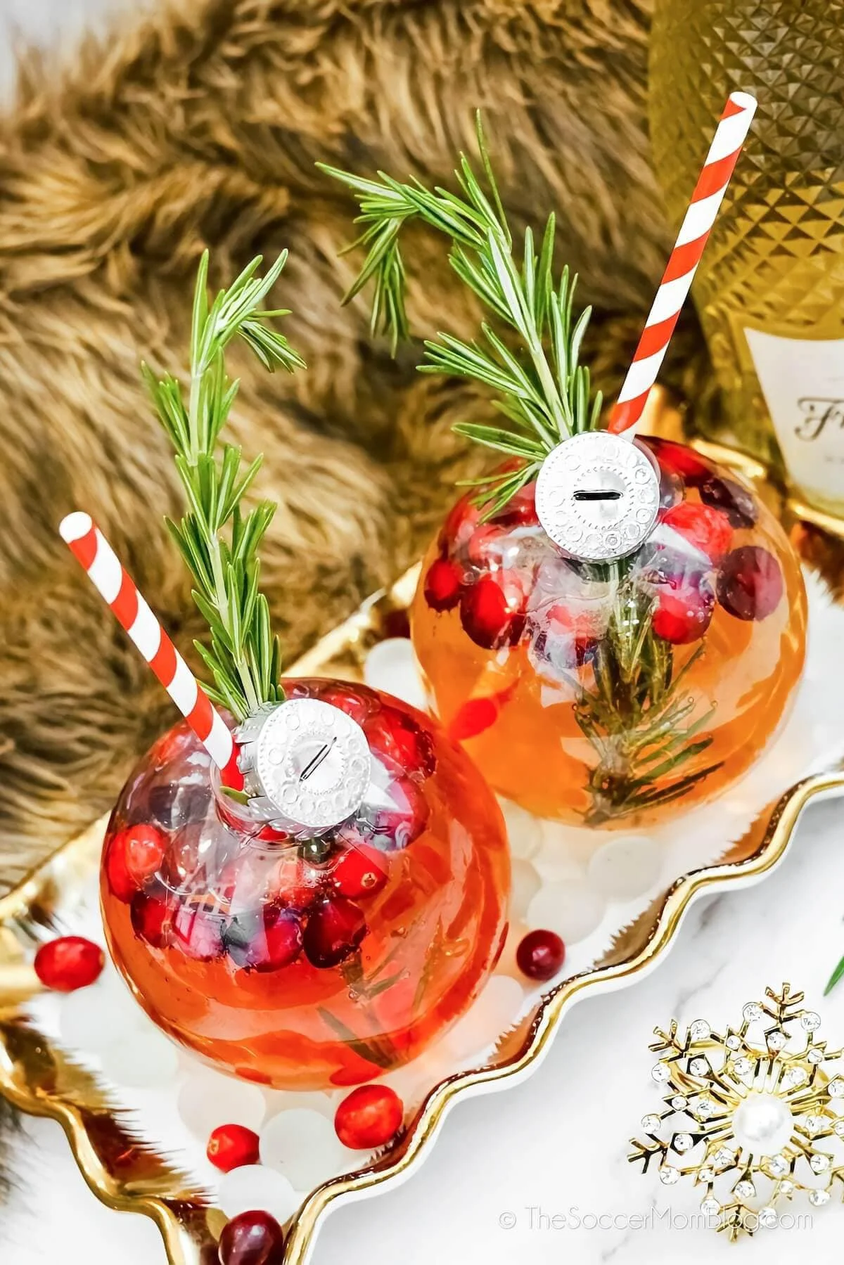 Decorative glass containers filled with red berries and sprigs of rosemary, adorned with striped straws and a silver medallion, placed on a white tray with a gold snowflake ornament on the side.
