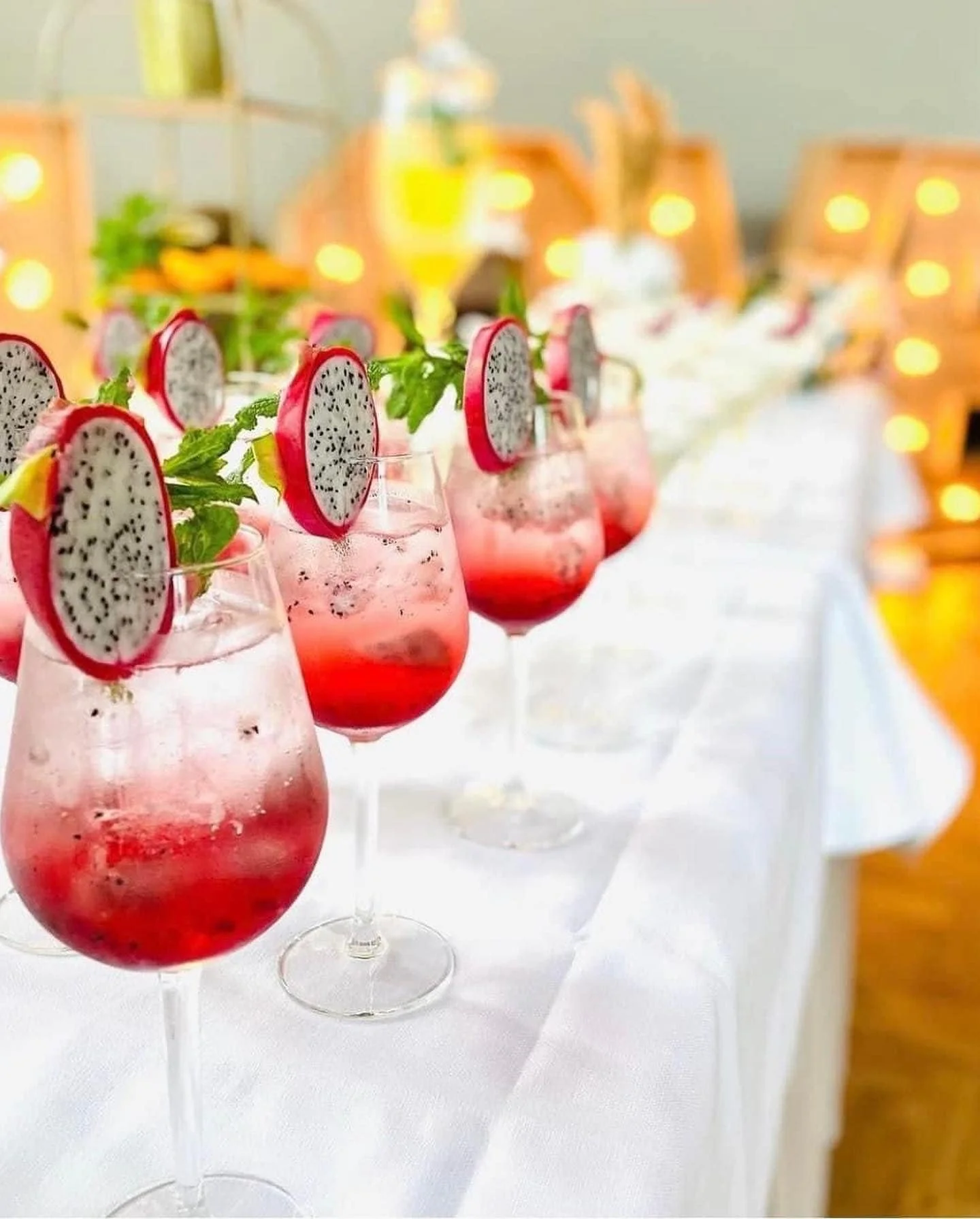 Row of pink cocktails garnished with dragon fruit slices and mint leaves on a white table at a festive event.