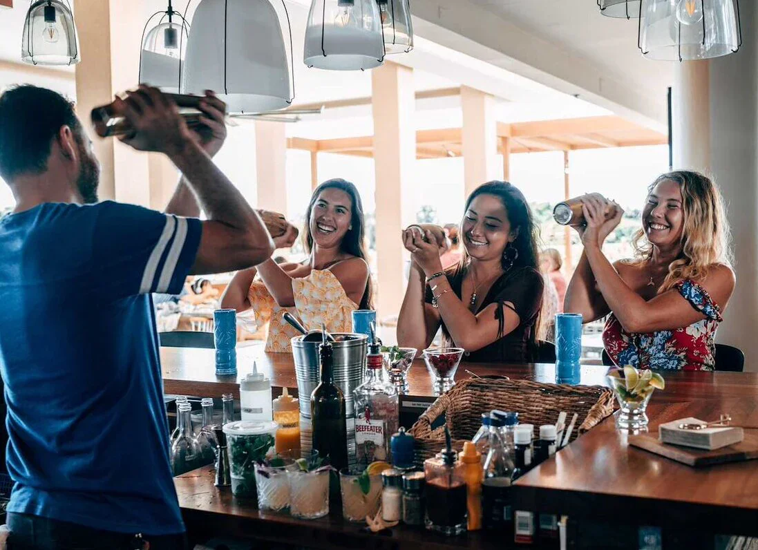 Four friends at a bar enjoying drinks and laughs, with a bartender preparing a cocktail in the background.