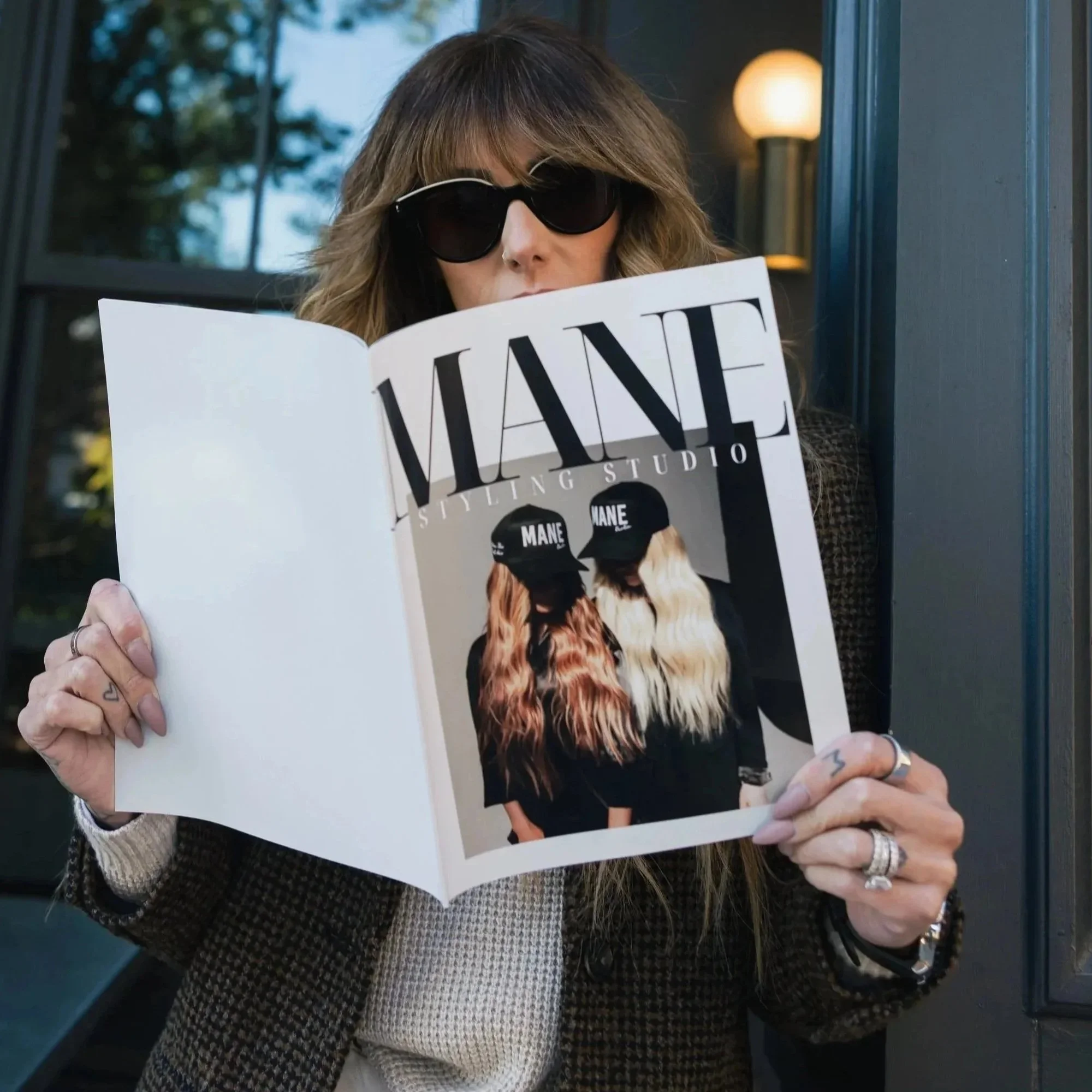 A woman with long wavy hair wearing sunglasses and a checkered jacket, holding a magazine that features two women with long hair and caps reading "MANE". The woman is standing outside near a building with a window and a lamp.