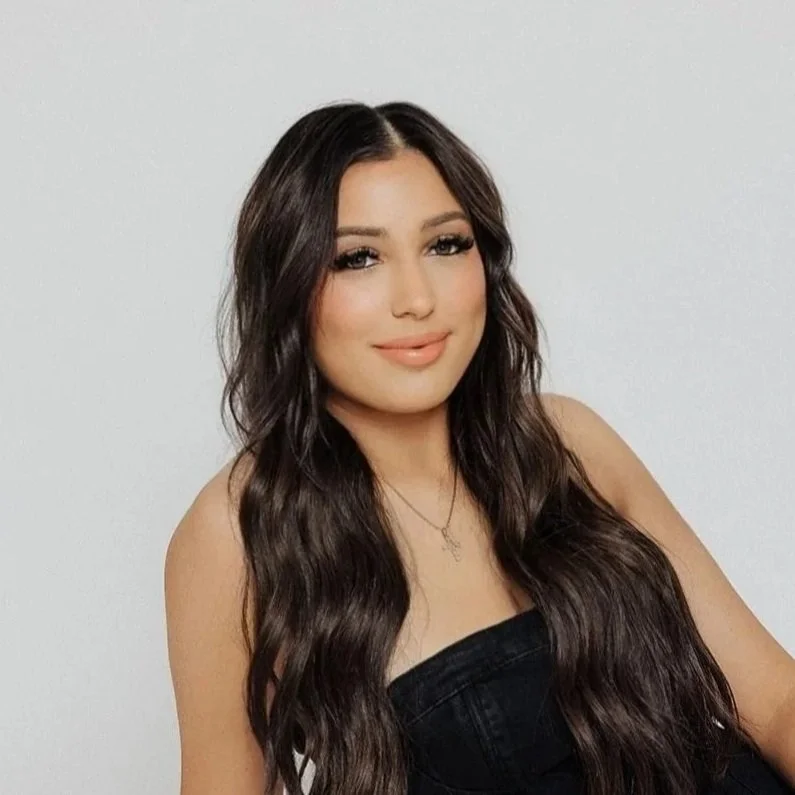 A woman with long, wavy dark brown hair, makeup, and a necklace, wearing a black strapless top, smiling at the camera against a plain white background.